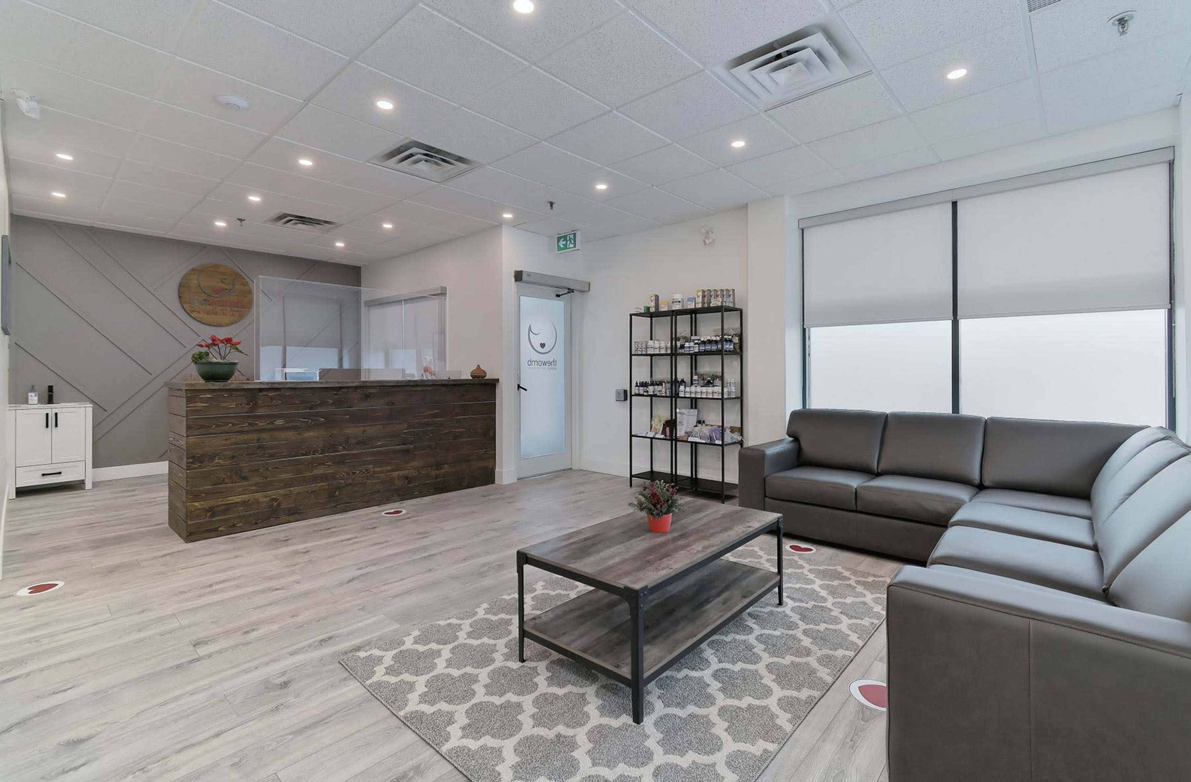 The image shows a modern reception area with a dark wood desk, gray seating, a coffee table, and a decorative carpet.