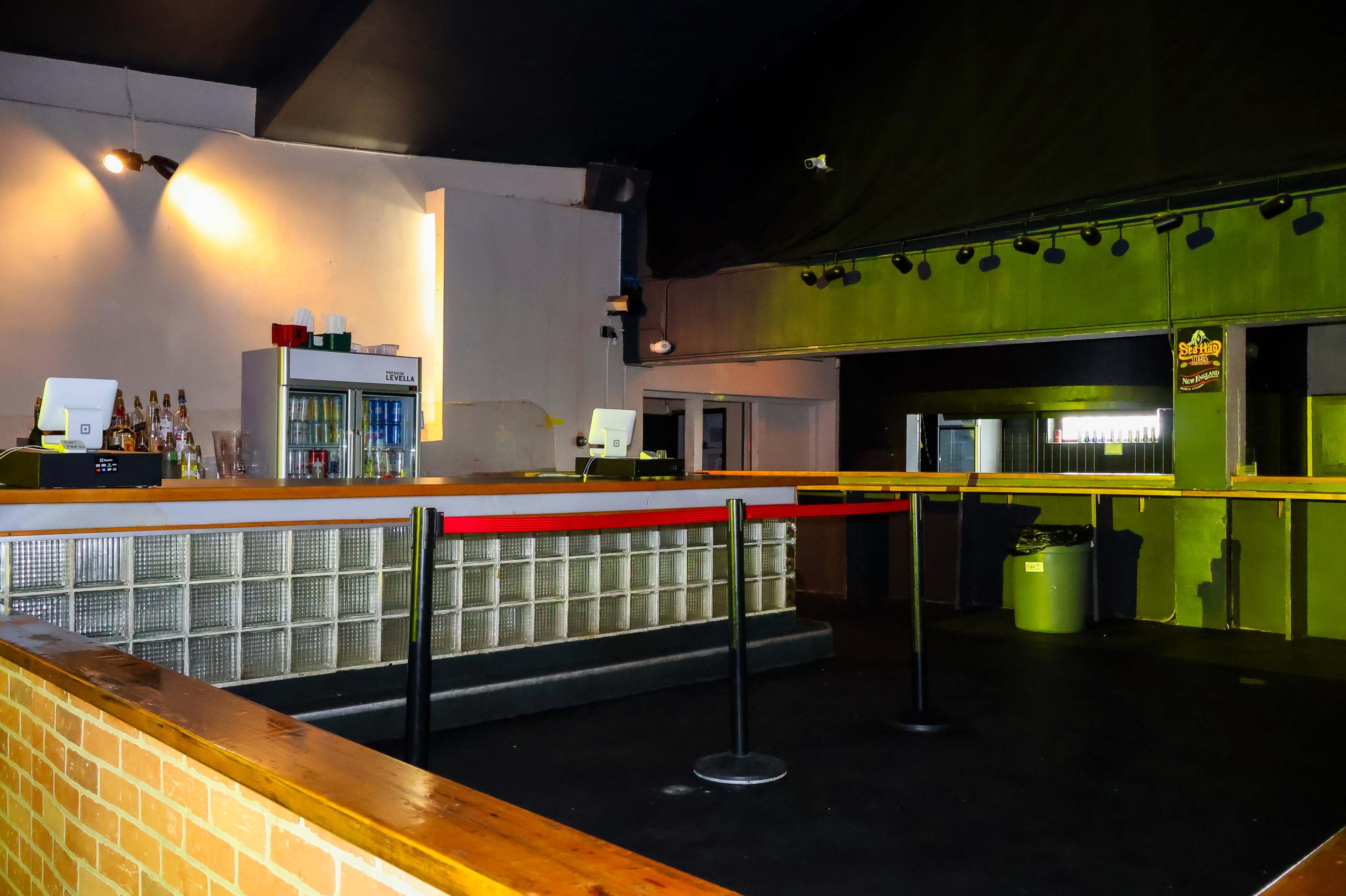 The image shows an empty bar area inside a dimly lit venue, featuring a counter, a beverage refrigerator, and stanchions with a red rope barrier.