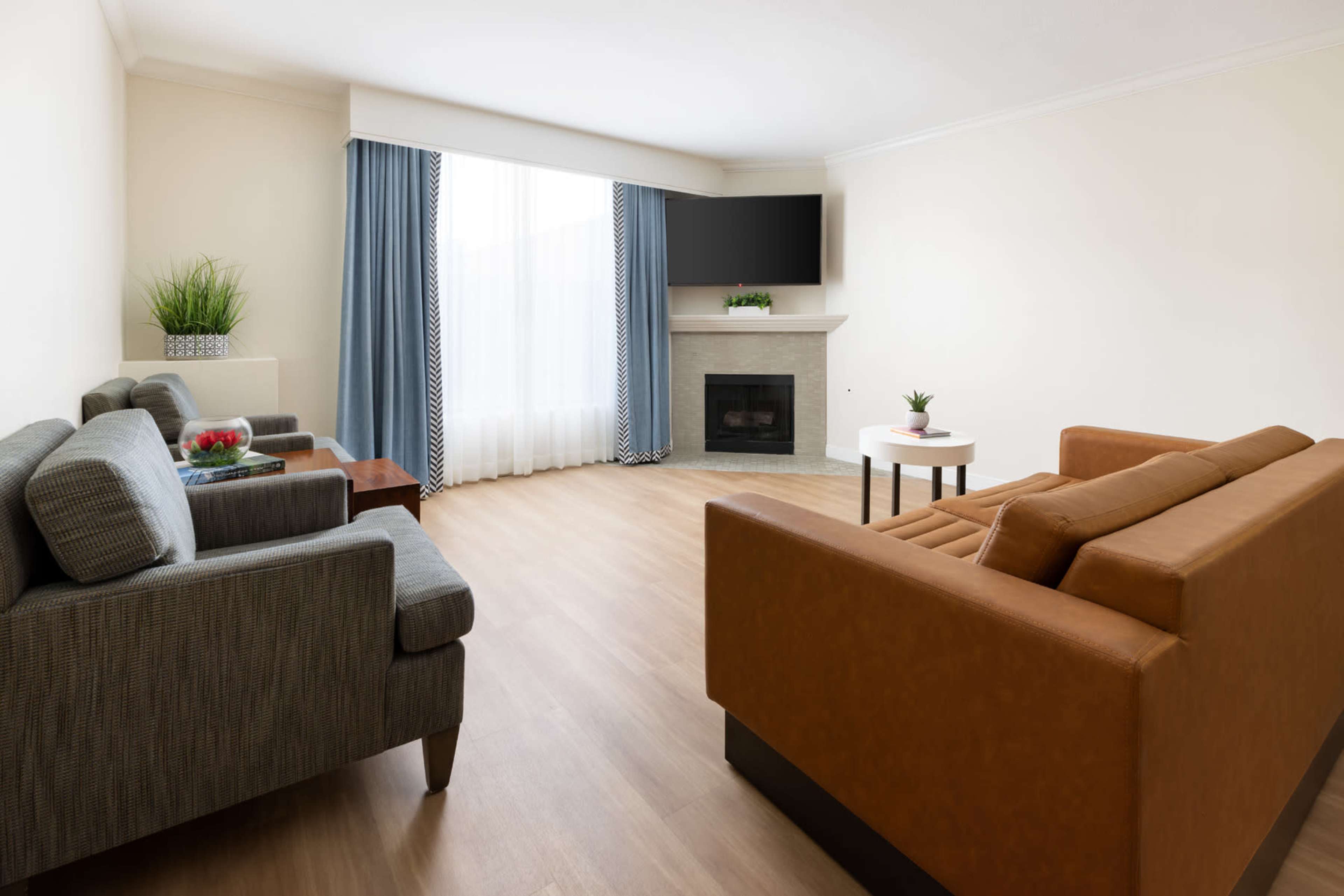 The image shows a modern living room with a brown sofa, gray armchair, a coffee table, a fireplace, and a television mounted above it, alongside large windows covered by light blue curtains.