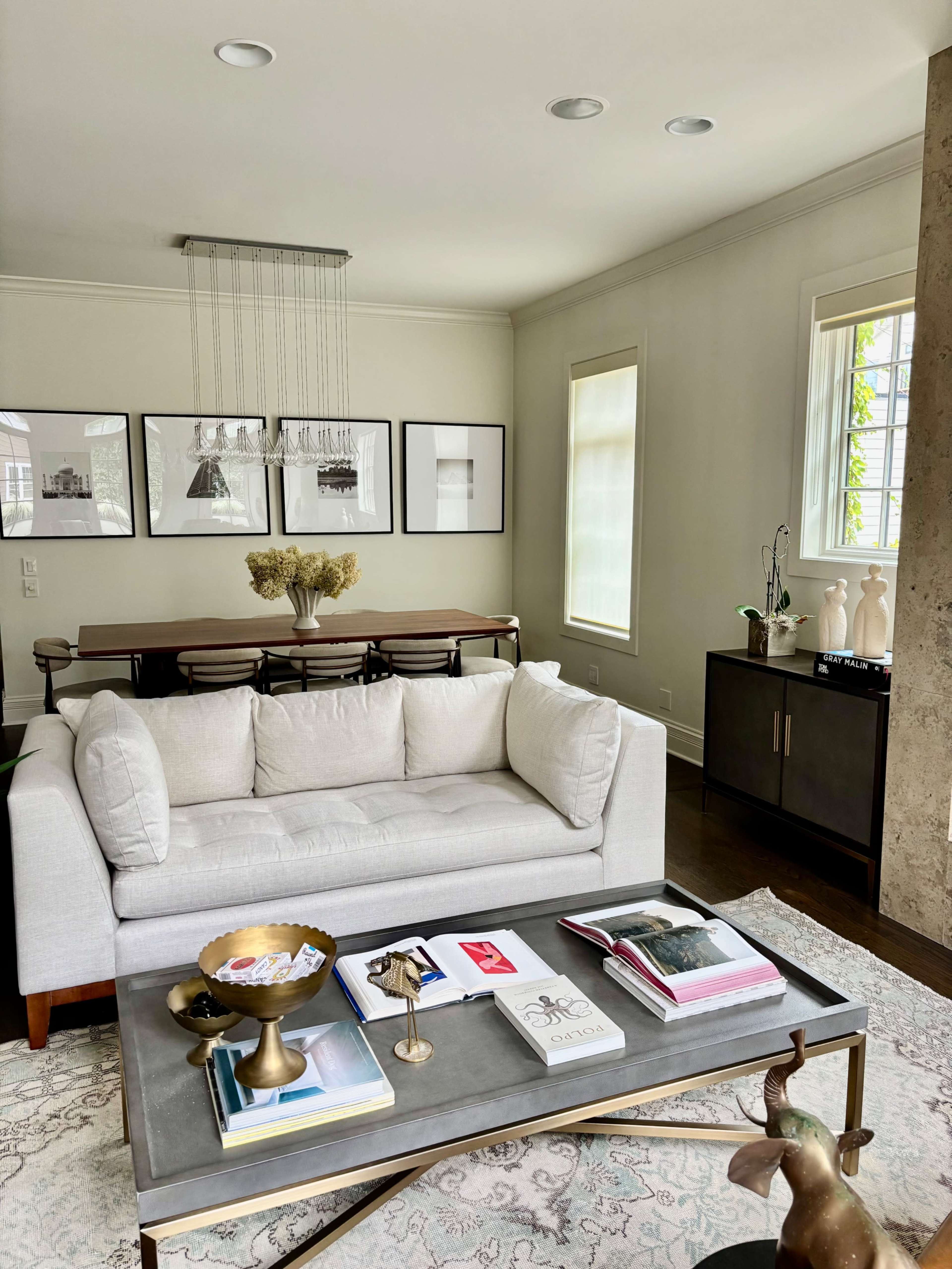 The image shows a modern living room featuring a light-colored sofa, a coffee table with books and decorative objects, and a dining area with a wooden table and framed artwork on the walls.