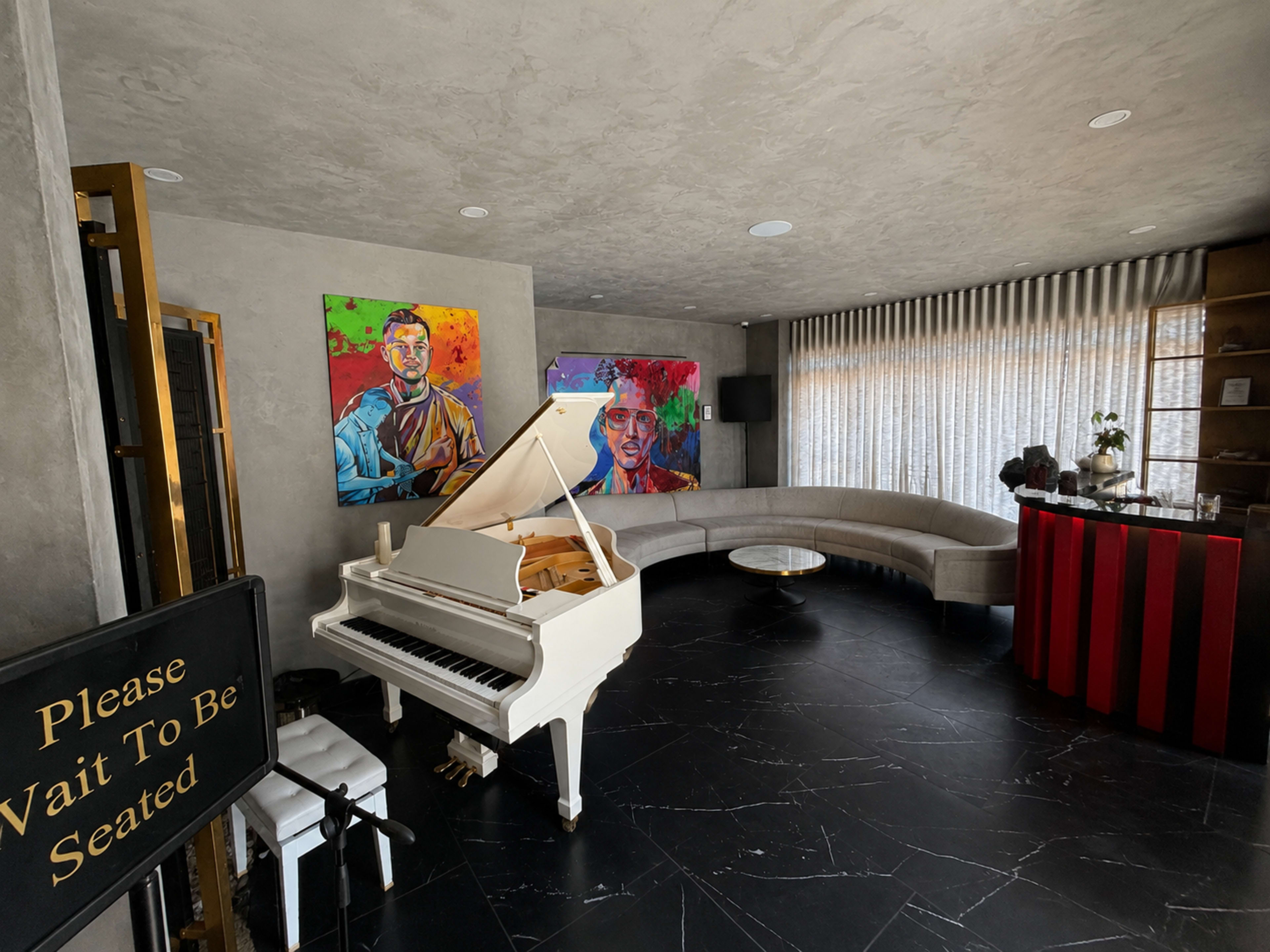 The image shows a modern interior space featuring a white grand piano, two colorful portraits on the wall, a curved sofa, and a small bar area with a red color scheme.