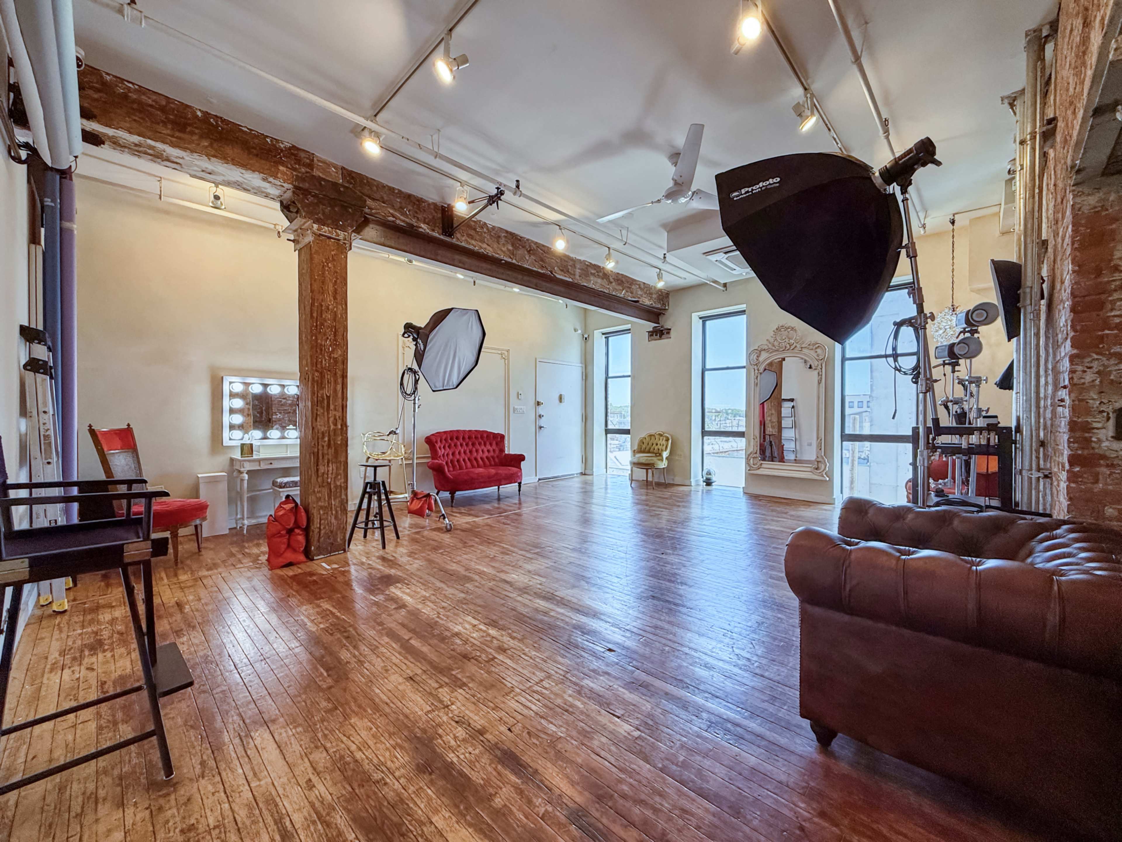 The image depicts a spacious studio featuring wooden floors, large windows, and photography equipment, along with a red velvet sofa and a vintage mirror.