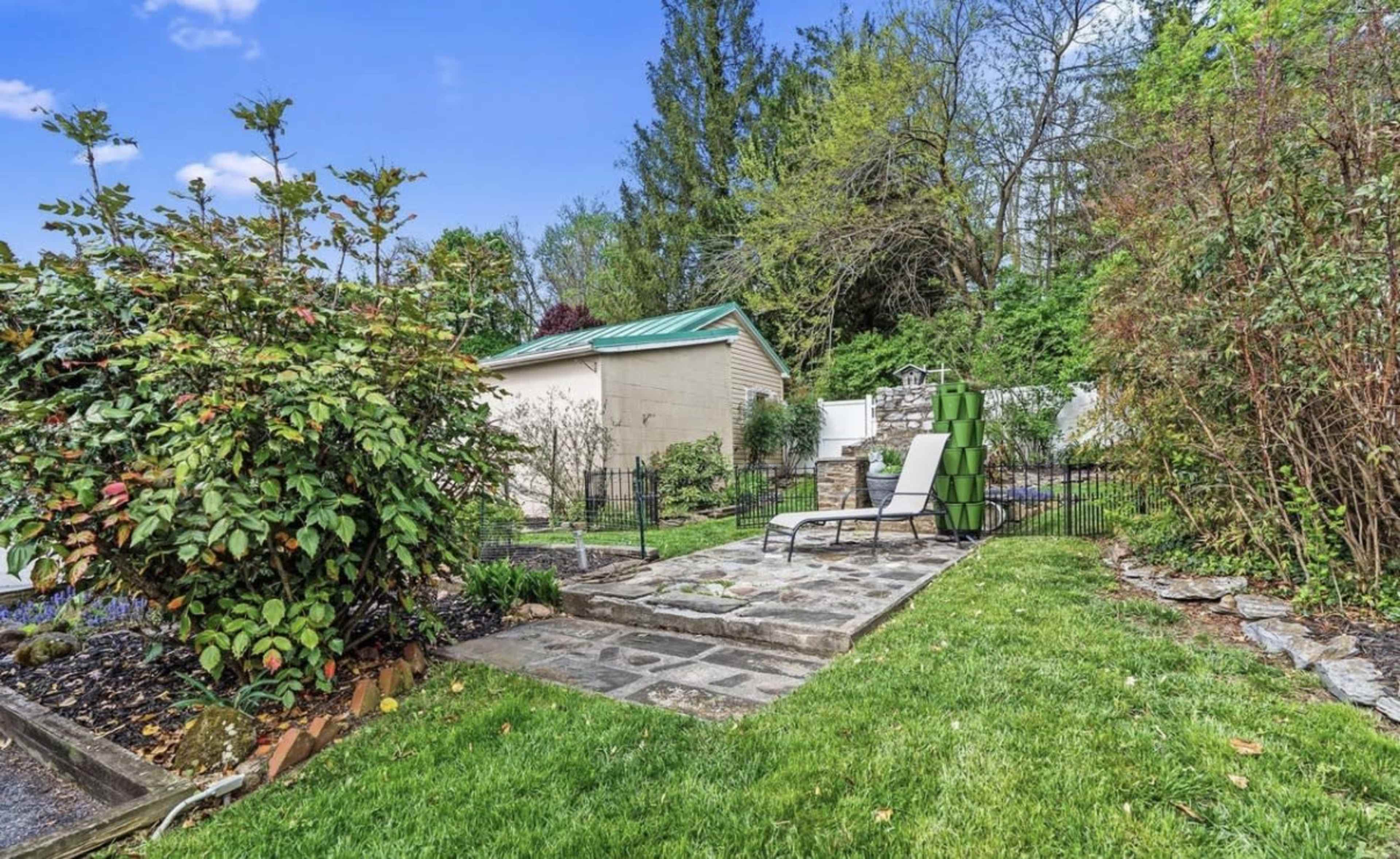 The image shows a landscaped outdoor space with a stone patio, a lounge chair, and a fenced area featuring greenery and a shed in the background.