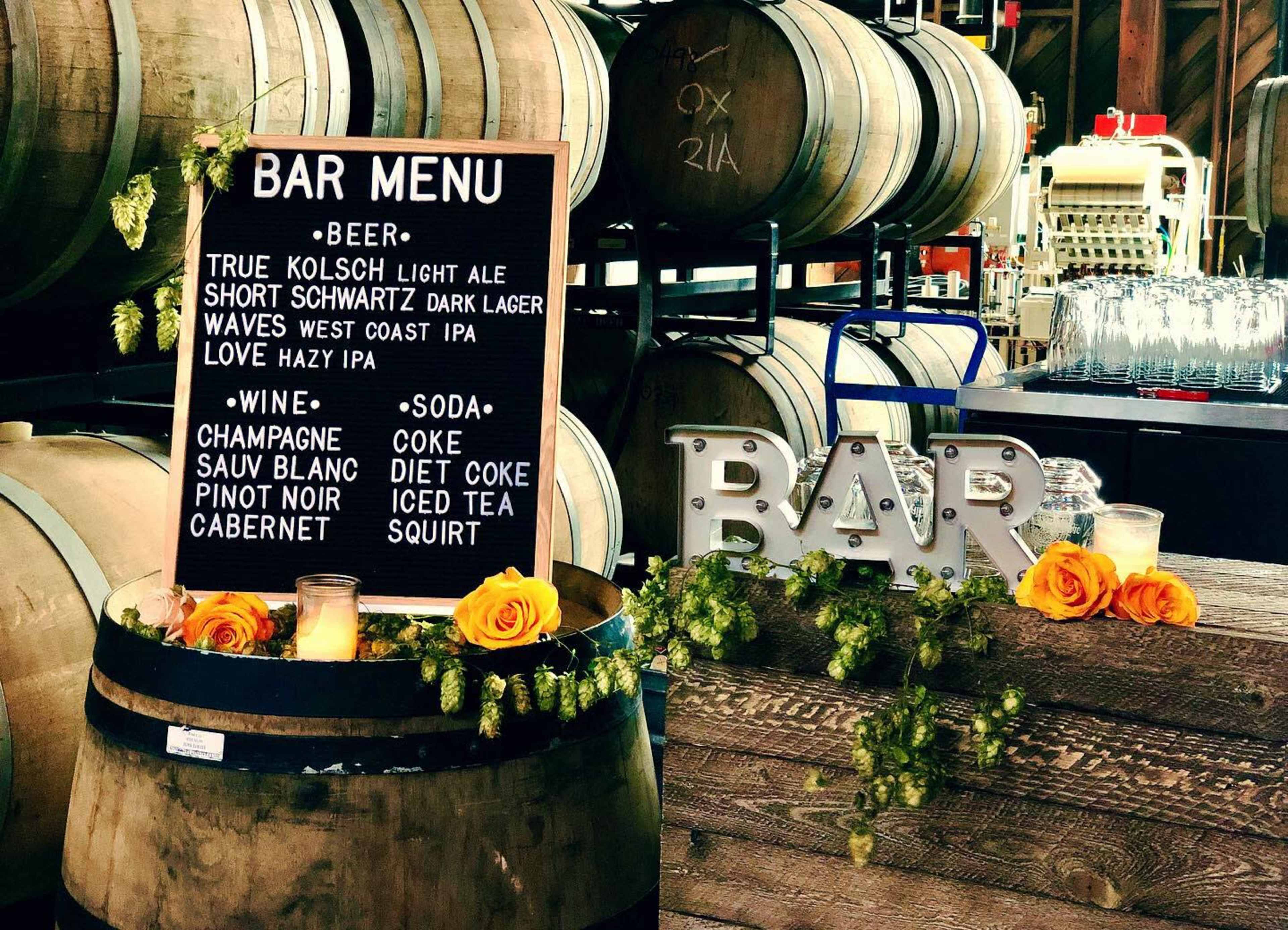 The image displays a bar menu board listing various beers, wines, and sodas, surrounded by wooden barrels and decorative flowers.