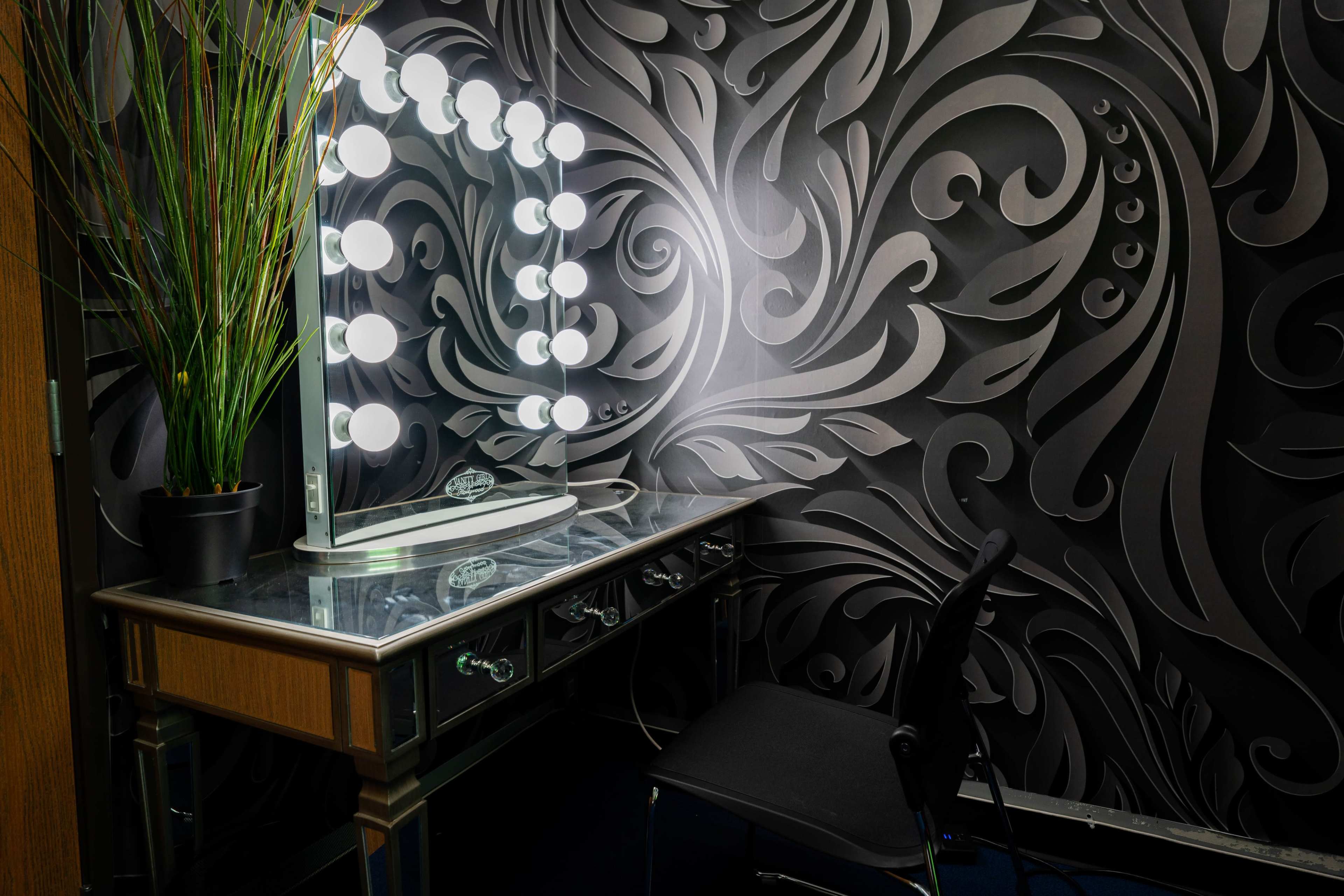 A dressing table with a large illuminated mirror is positioned against a patterned black wall.