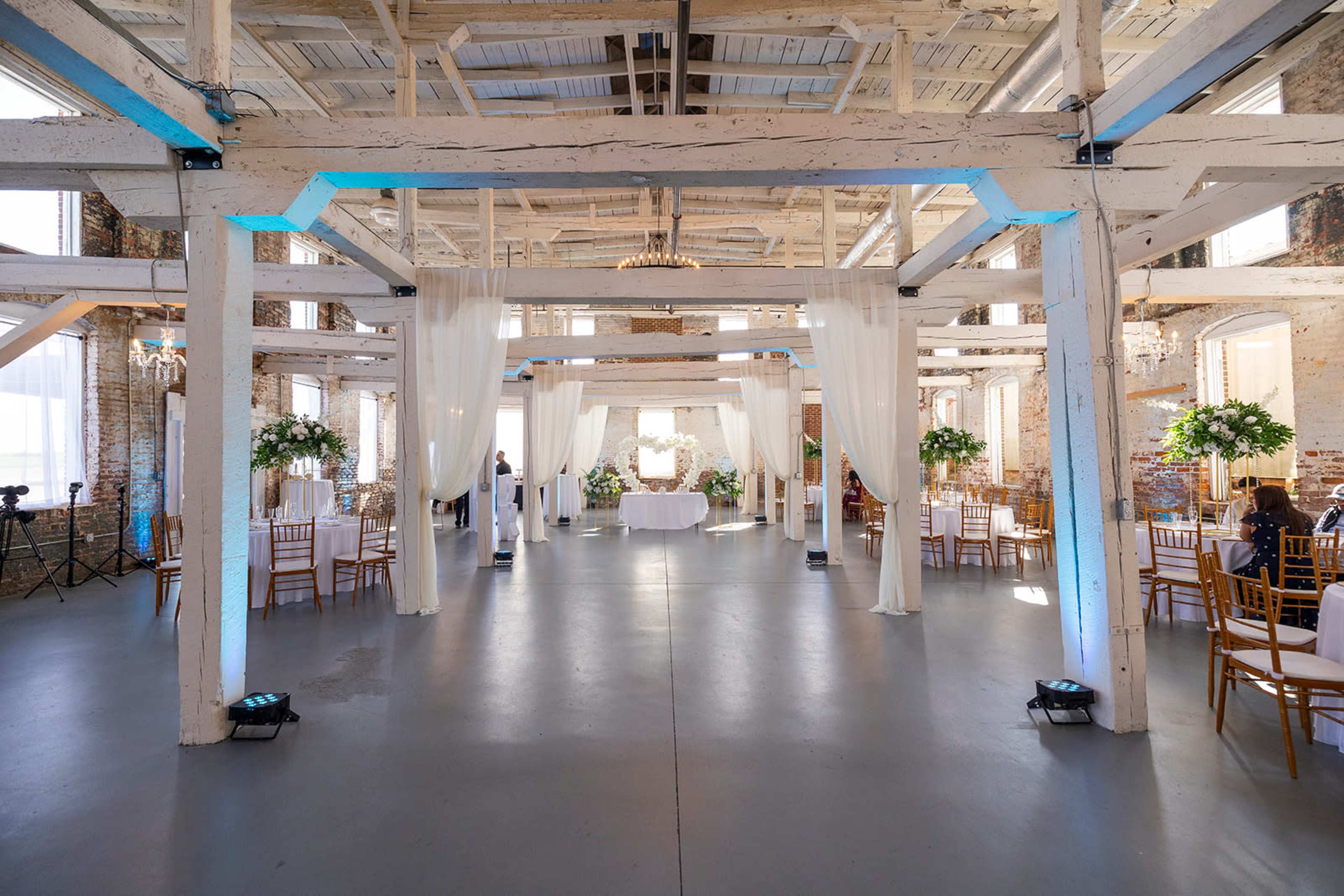 The scene shows a spacious, elegantly decorated venue with exposed wooden beams, draped white fabric, and floral arrangements, set for an event with tables and chairs arranged throughout.