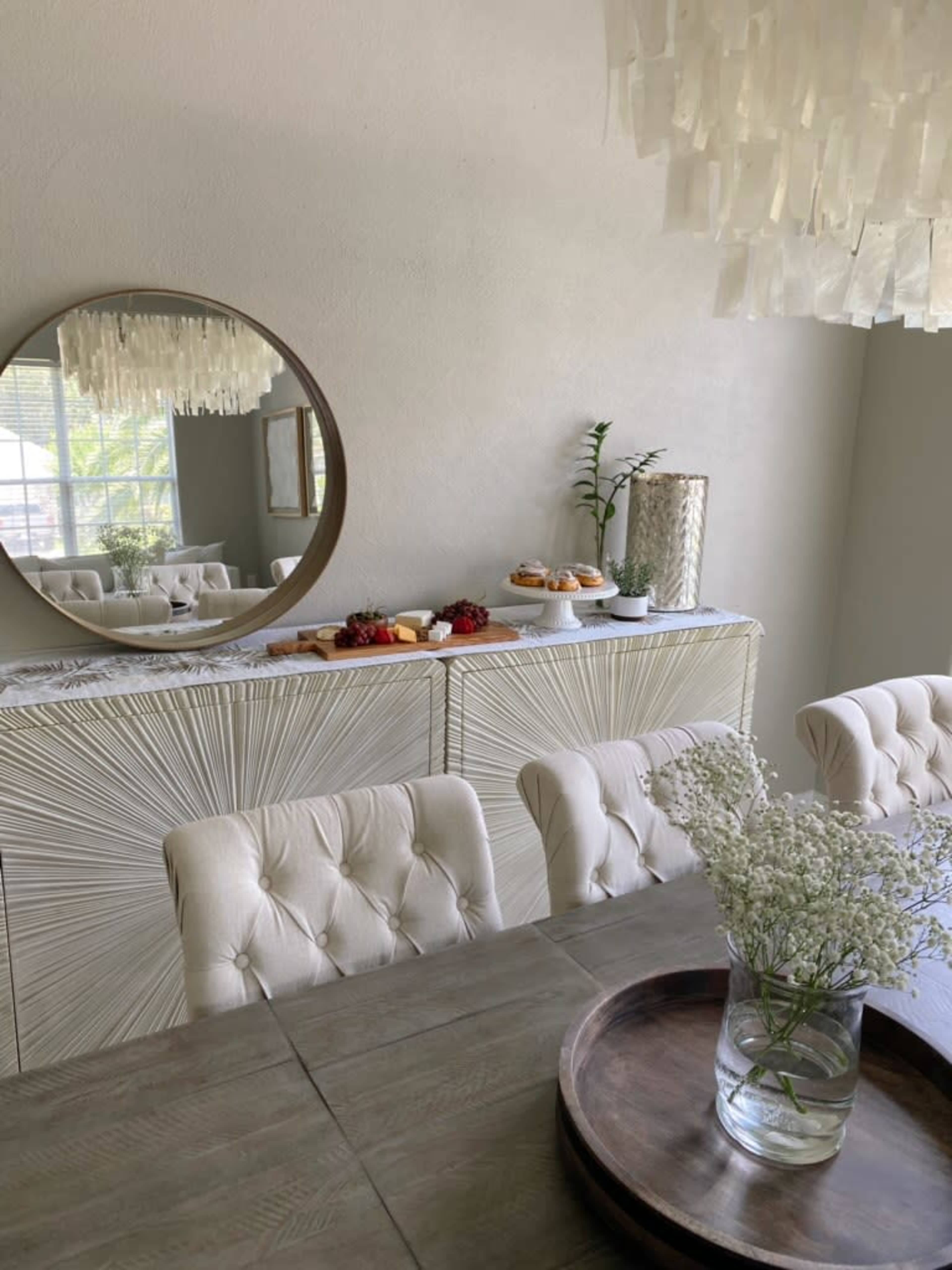 A dining area features a round mirror above a sideboard decorated with pastries and a plant, accompanied by a wooden table set with a vase of flowers.