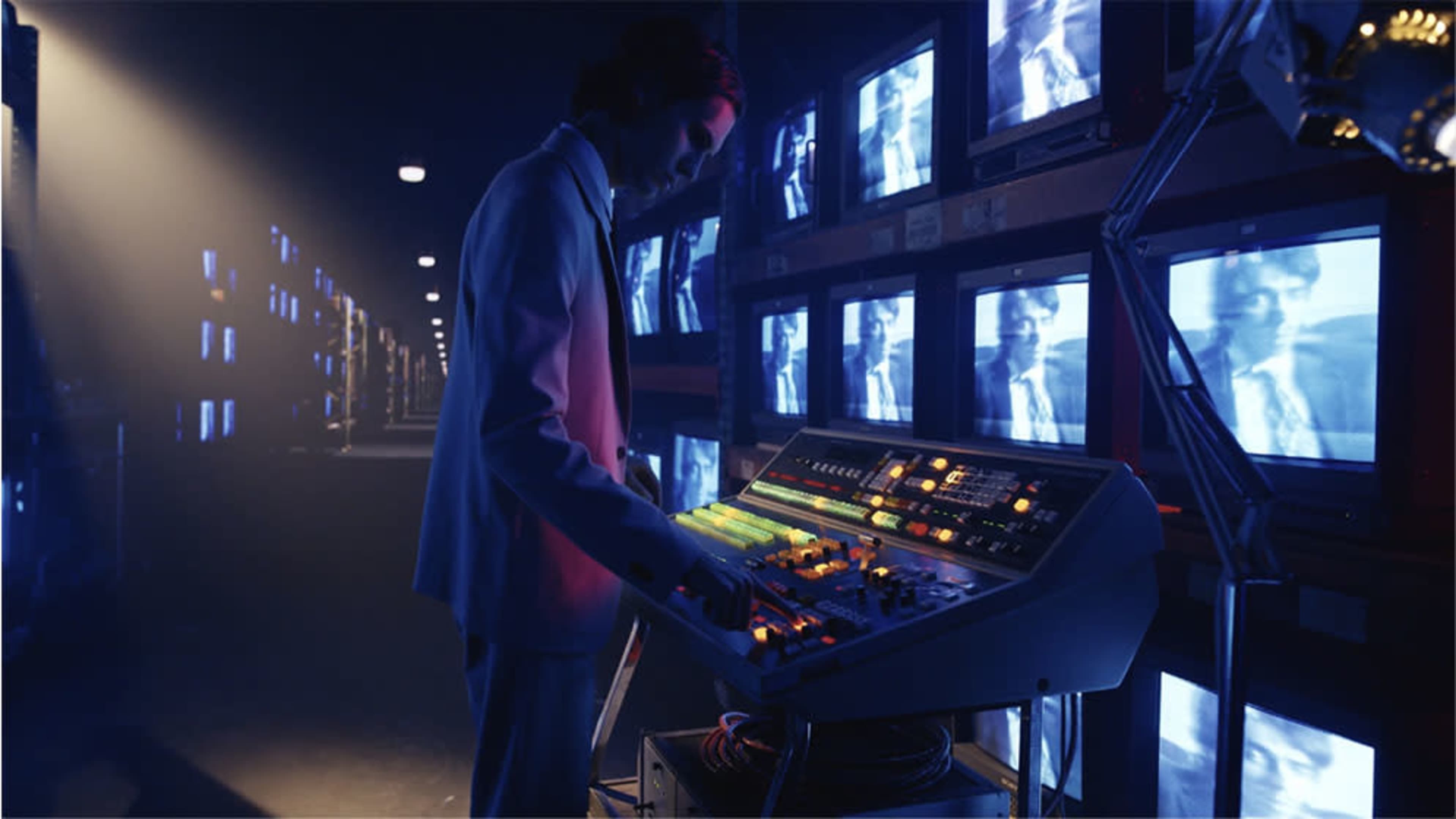 A person in a suit operates a colorful control panel in a dimly lit room filled with flickering television screens.