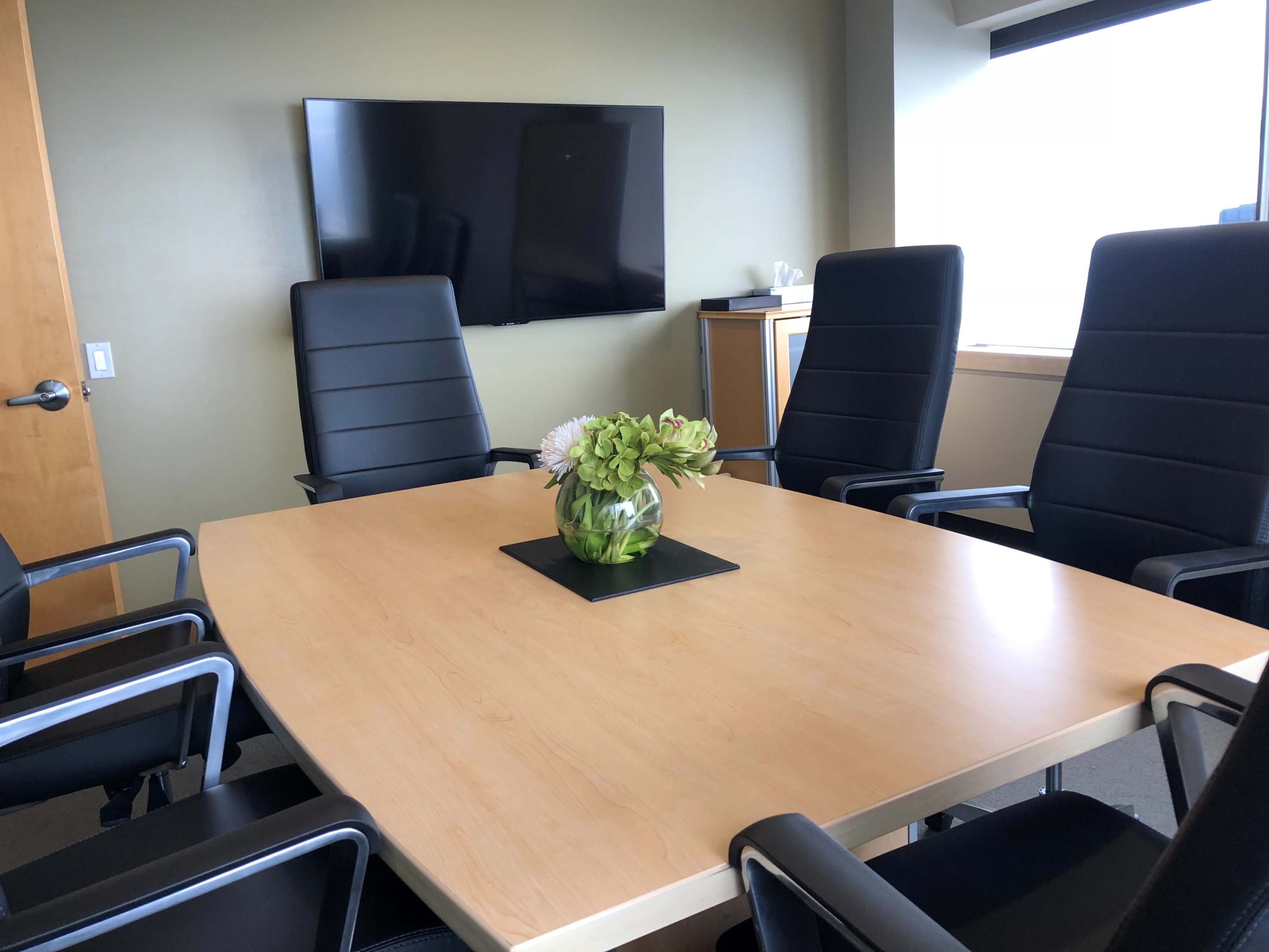A conference room features a large wooden table surrounded by six black chairs, with a television mounted on the wall and a floral arrangement in the center.
