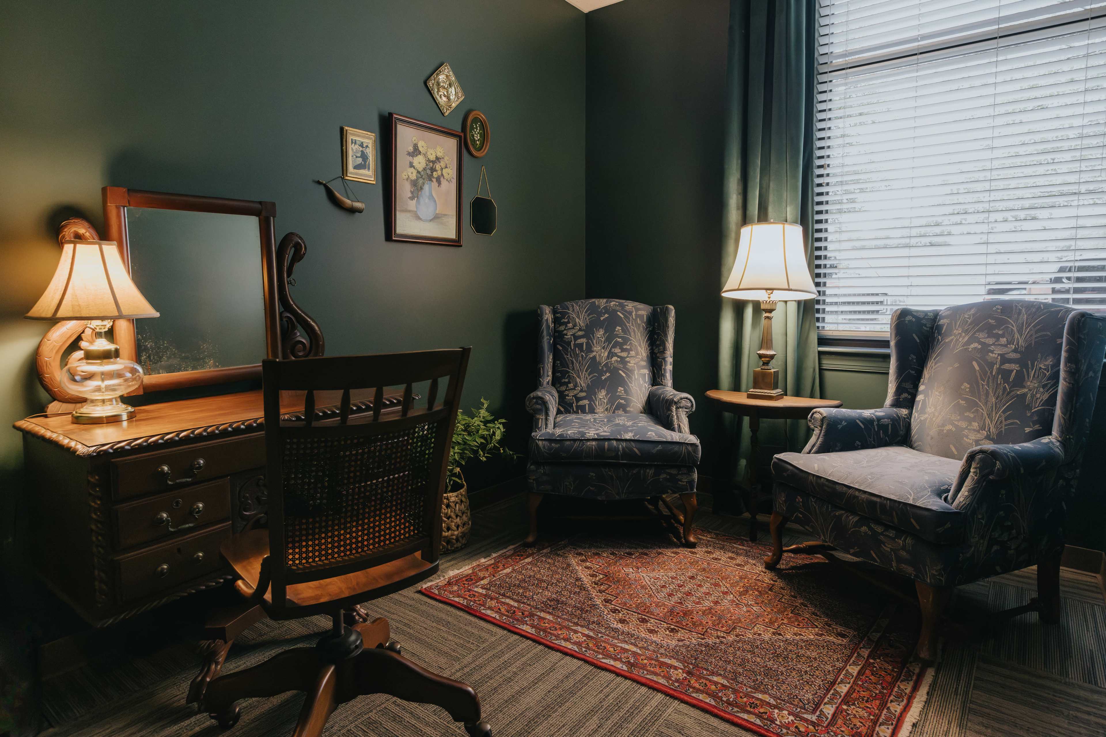 A cozy corner of a room featuring two upholstered chairs, a wooden desk with a mirror, a small side table with a lamp, and a patterned rug.