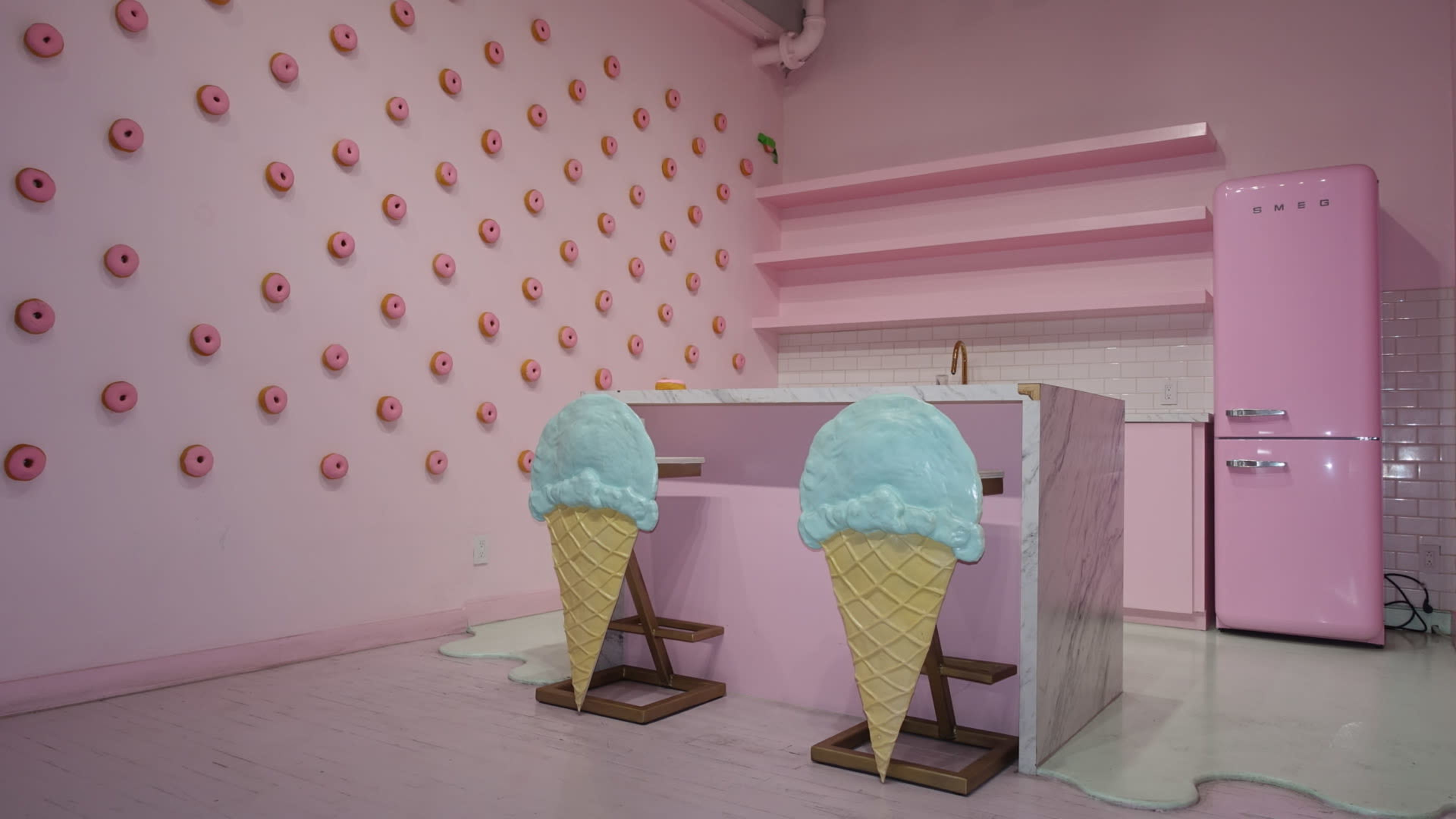 The image features a brightly colored pink room with a wall adorned with donut decorations and a central table designed to resemble ice cream cones.