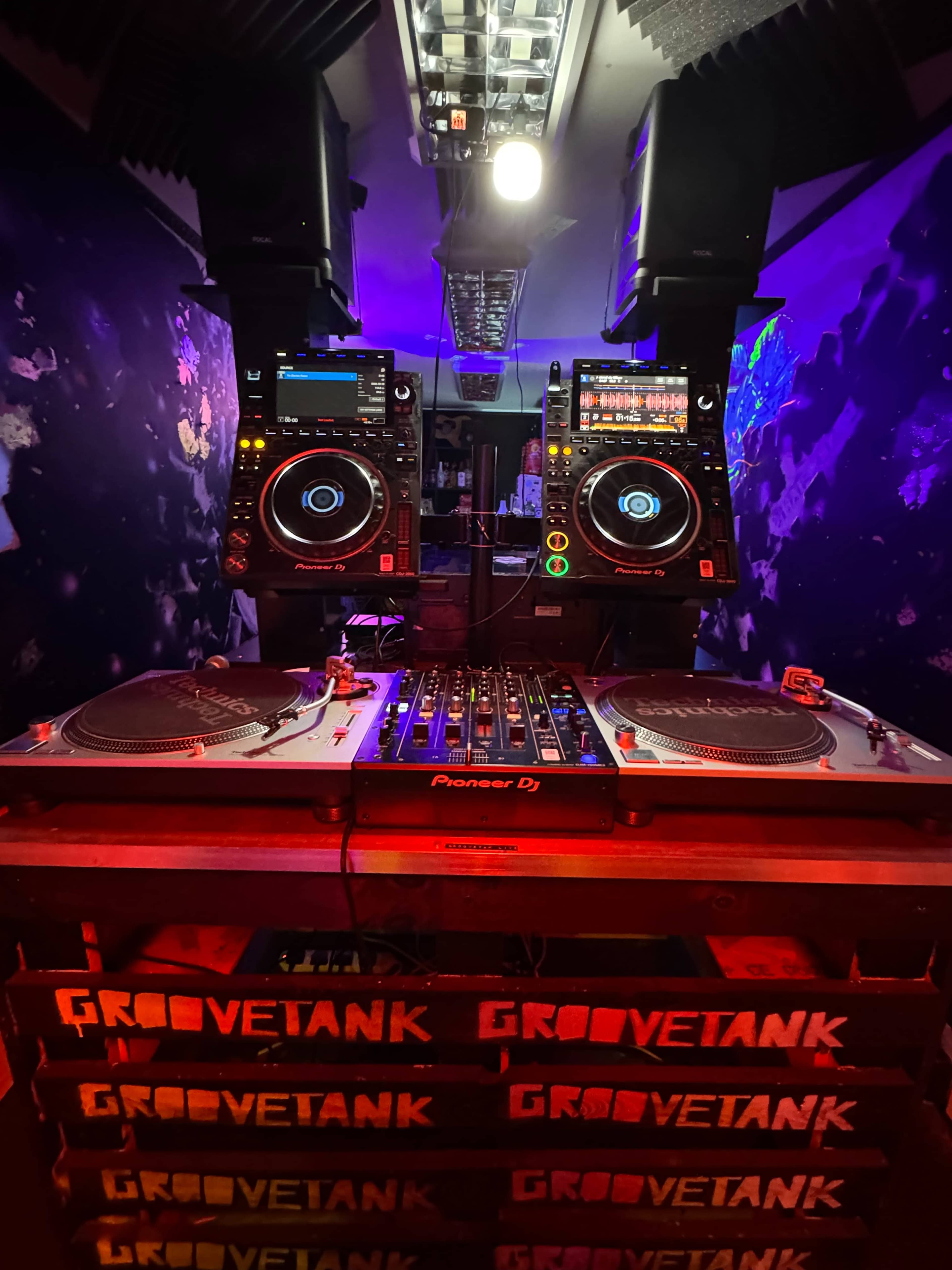 A DJ setup features two turntables and a mixer, with equipment illuminated by purple and blue lights in a dimly lit room.