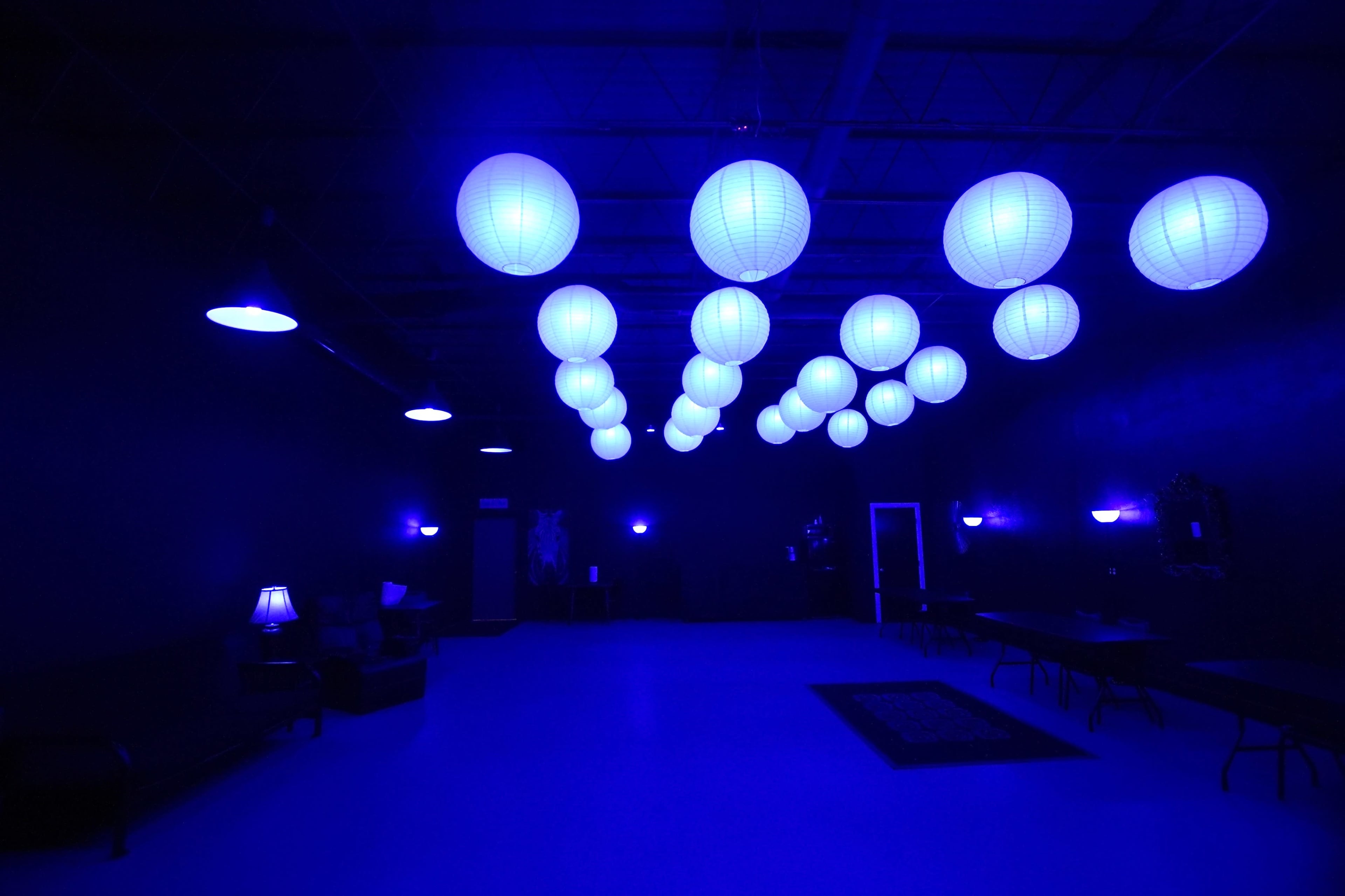 A dimly lit space with blue paper lanterns hanging from the ceiling and minimal furnishings, including a few chairs and tables.
