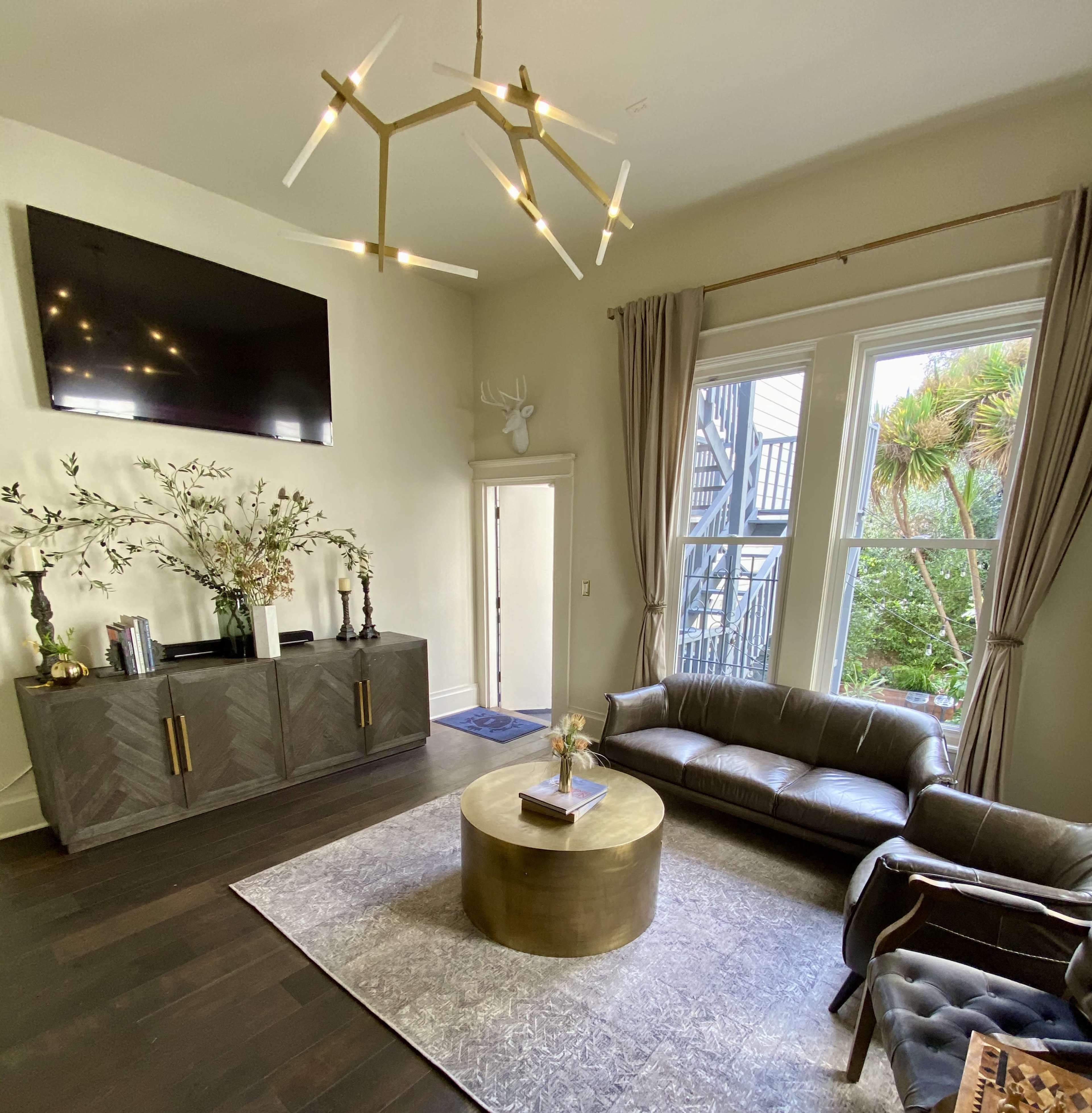 The living room features a modern design with a television mounted on the wall, a stylish coffee table, large windows, and a sofa, alongside decorative plants and accessories.