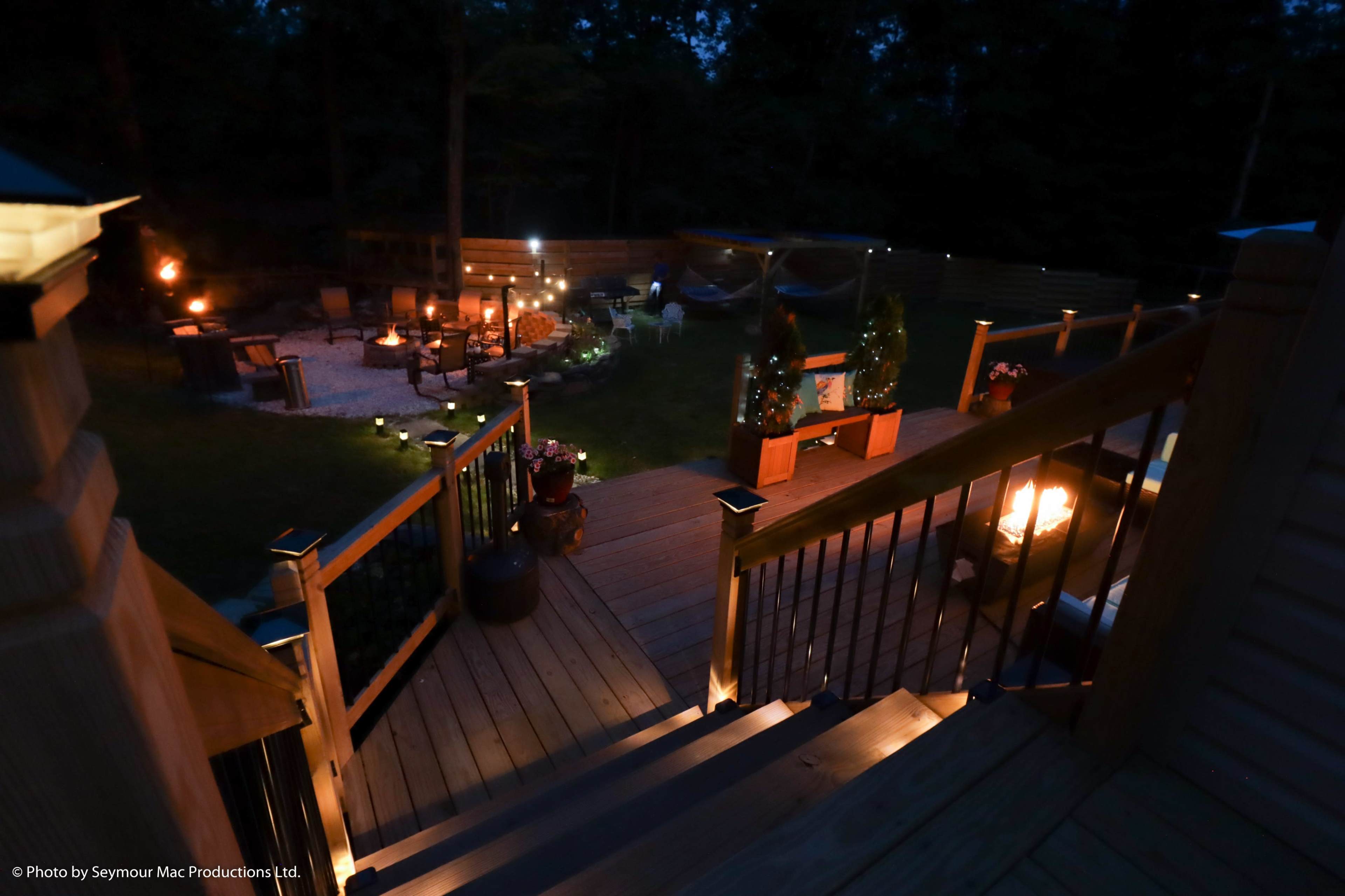 The image shows a wooden deck with a staircase leading to a garden area illuminated by soft lights, featuring seating arrangements around a fire pit and decorative planters.