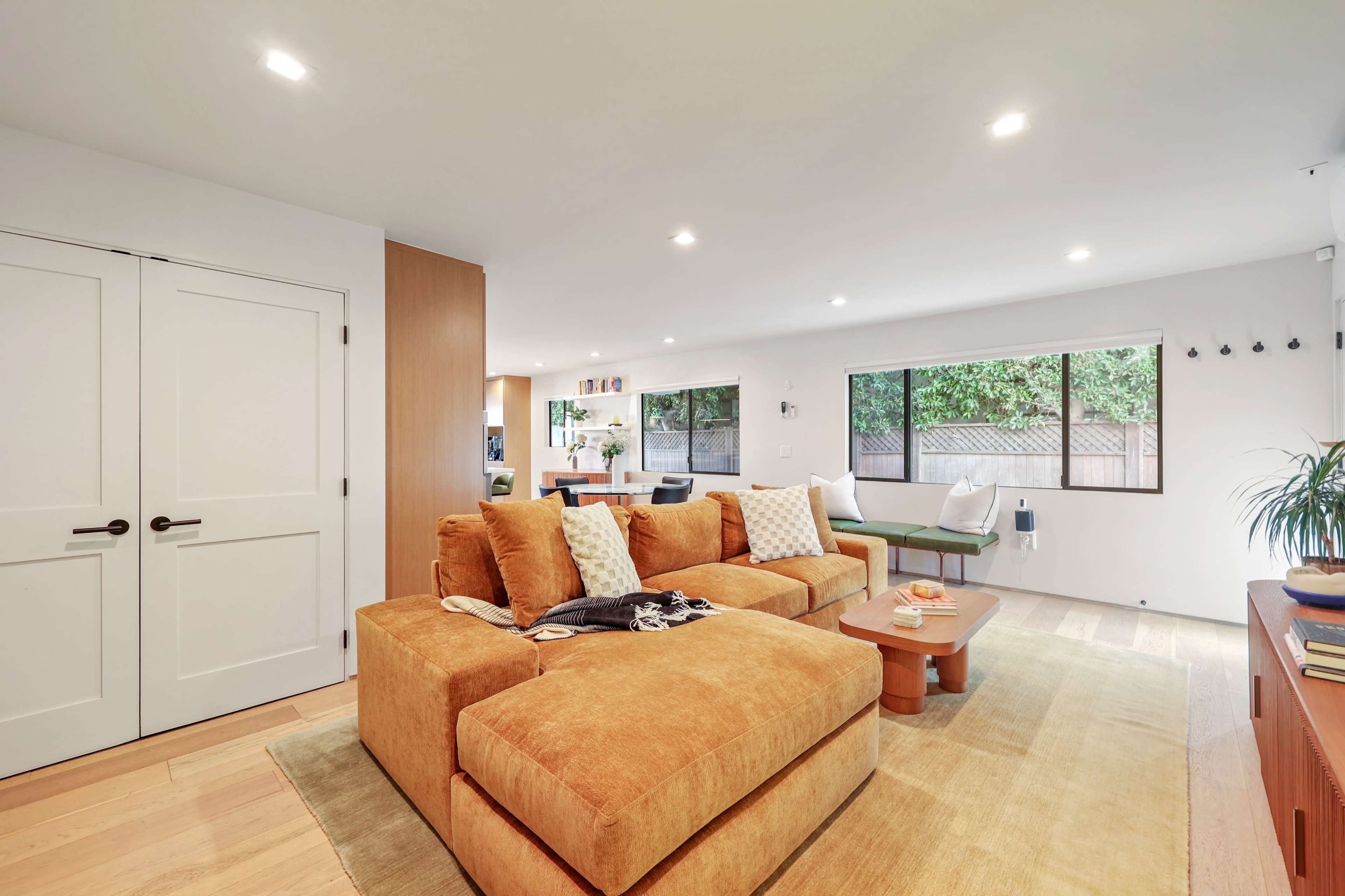 The image shows a modern living room with a large orange sectional sofa, wooden floor, and large windows allowing natural light.