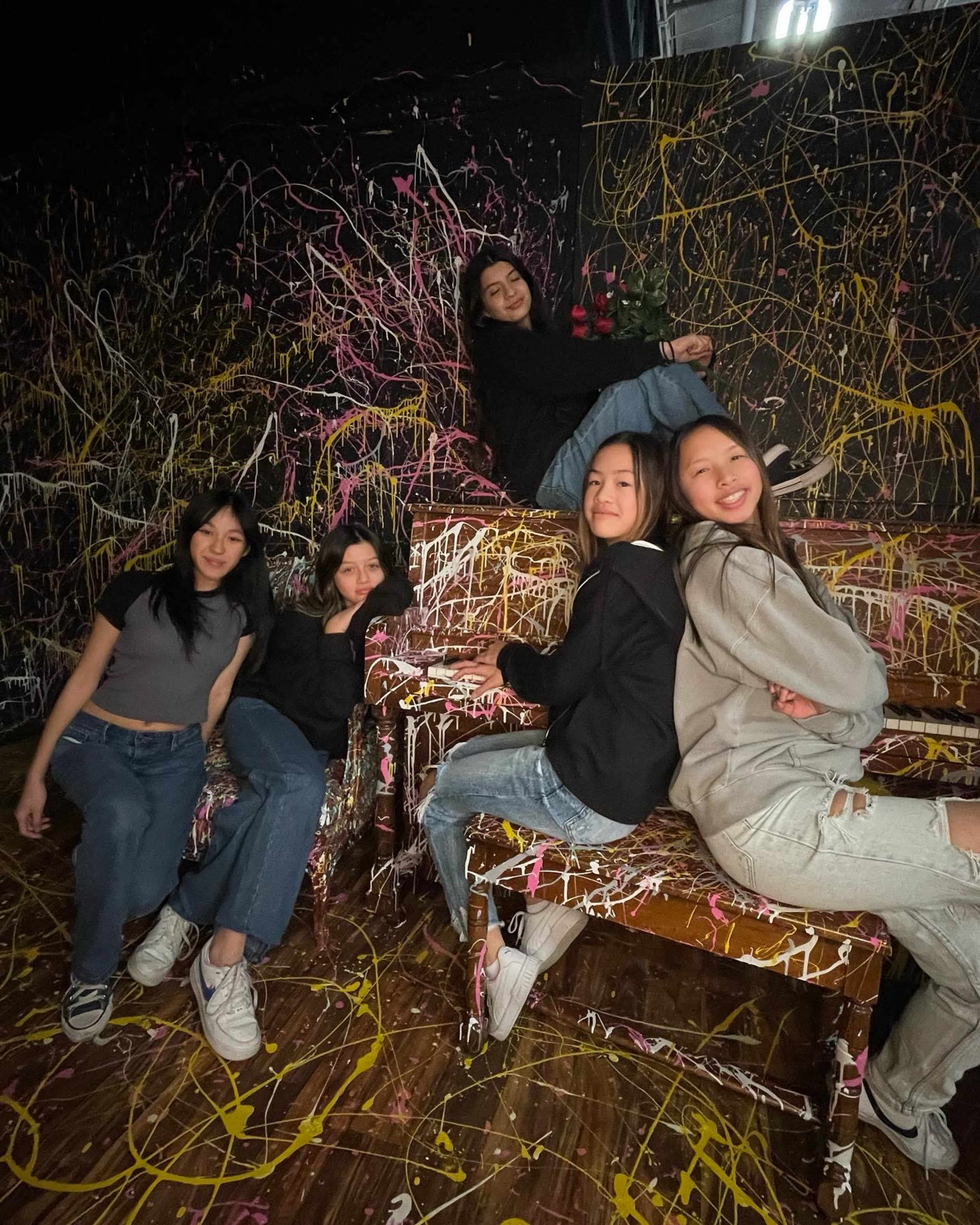 Five individuals pose on colorful, paint-splattered furniture in a vibrant room adorned with abstract paint splatter.
