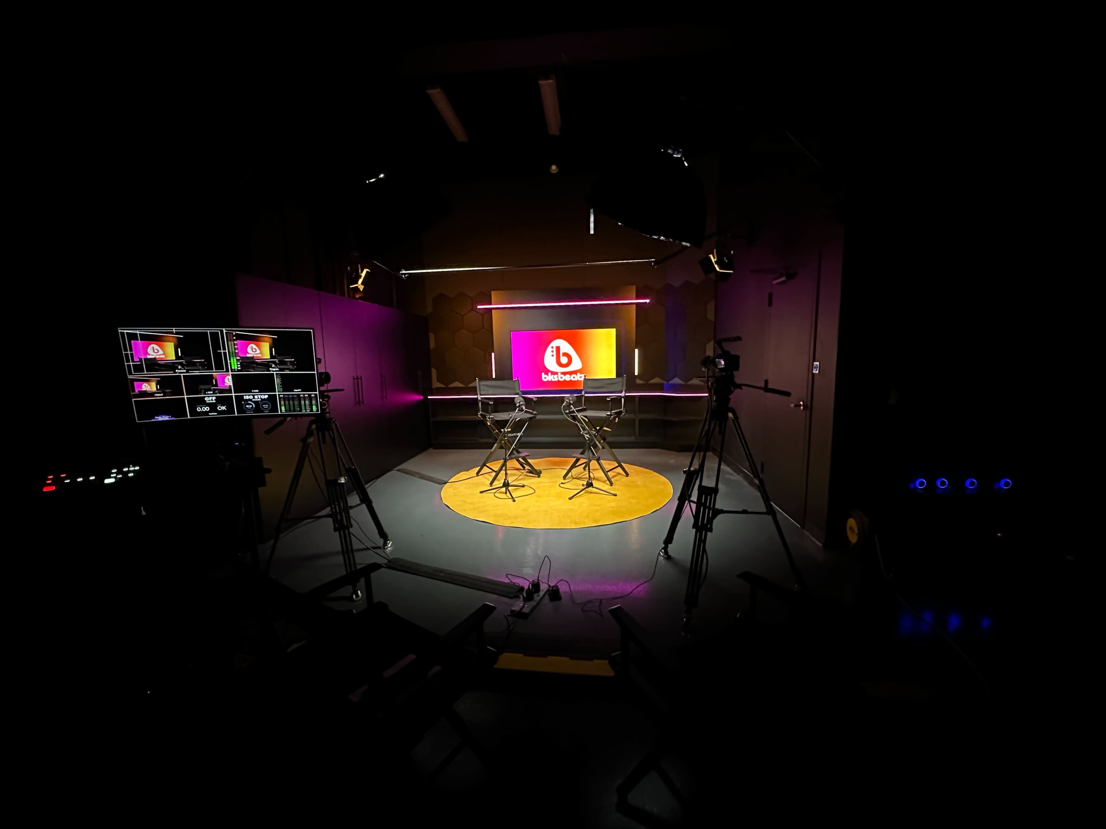 The image shows a television studio setup with two chairs in the center, surrounded by cameras and a large screen displaying a logo.