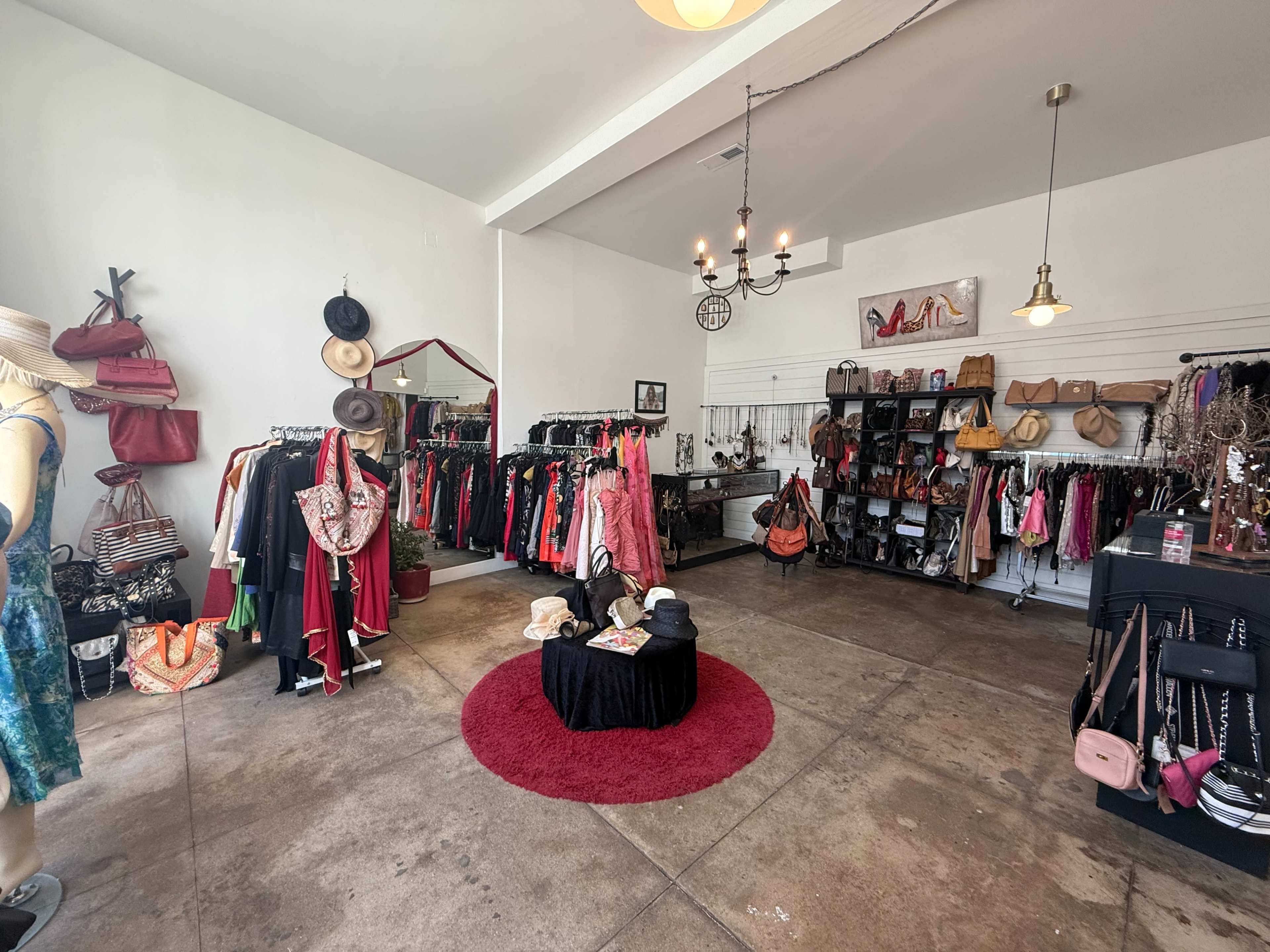 The image shows a boutique interior with clothing racks displaying various garments and accessories, a round red rug in the center, and hats on a mannequin.
