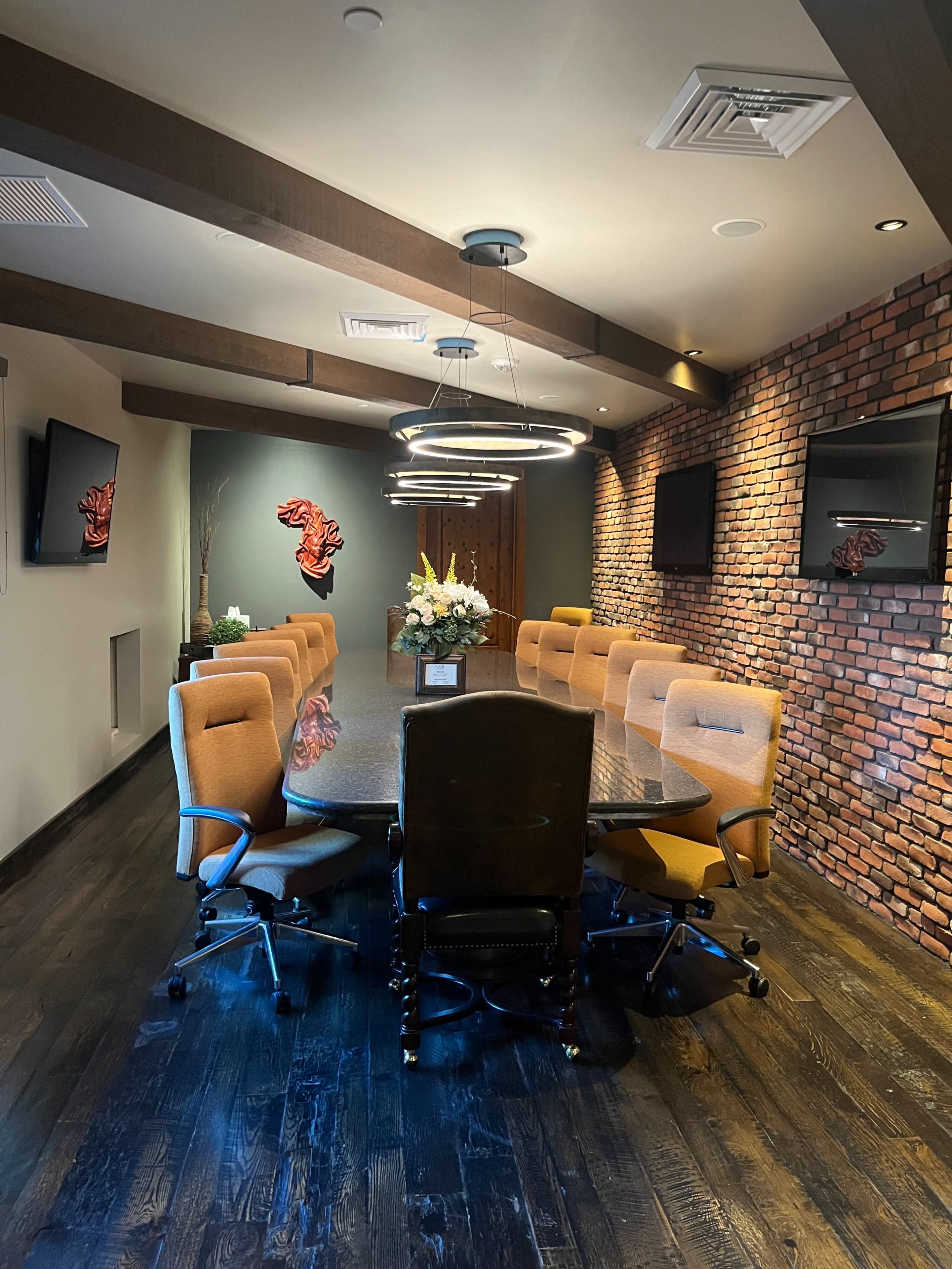 A conference room features a large circular table surrounded by orange chairs, accented by a brick wall and modern lighting fixtures.