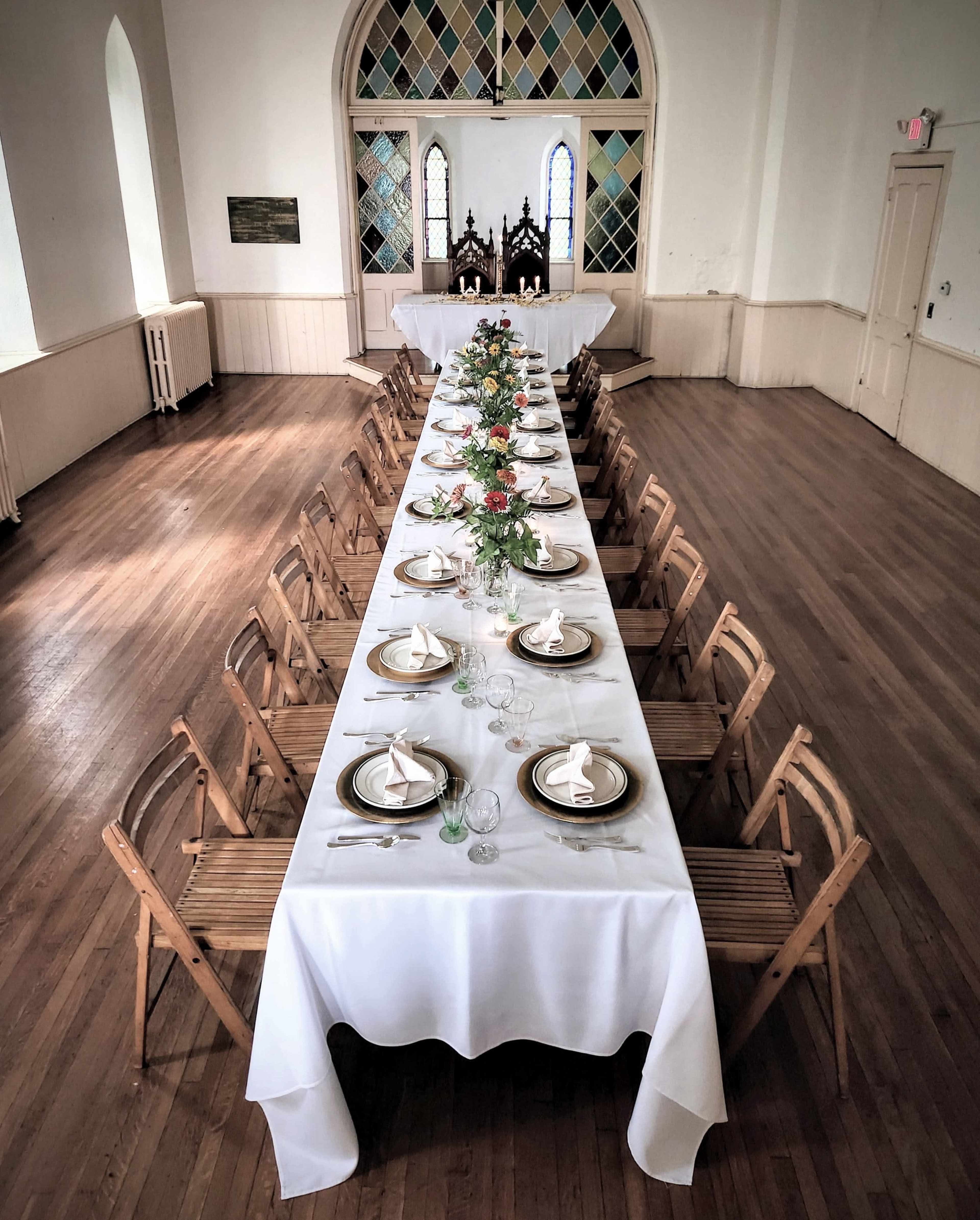 A long dining table is set with white tablecloths, plates, and floral centerpieces in a spacious, well-lit room with stained glass windows.