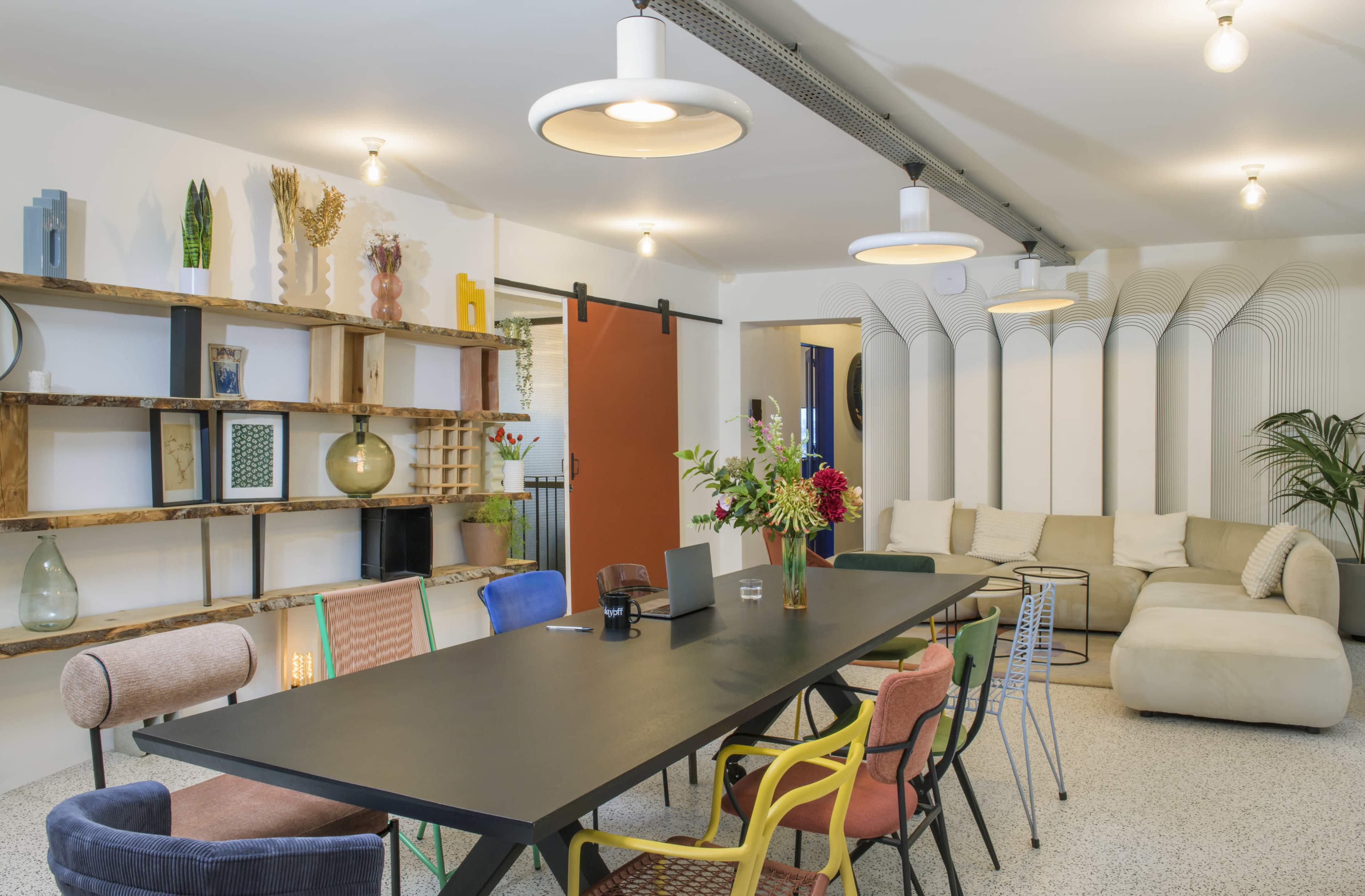 The image shows a modern, open-concept living space with a long black dining table surrounded by colorful chairs, a beige sectional sofa, and decorative shelves on the wall.