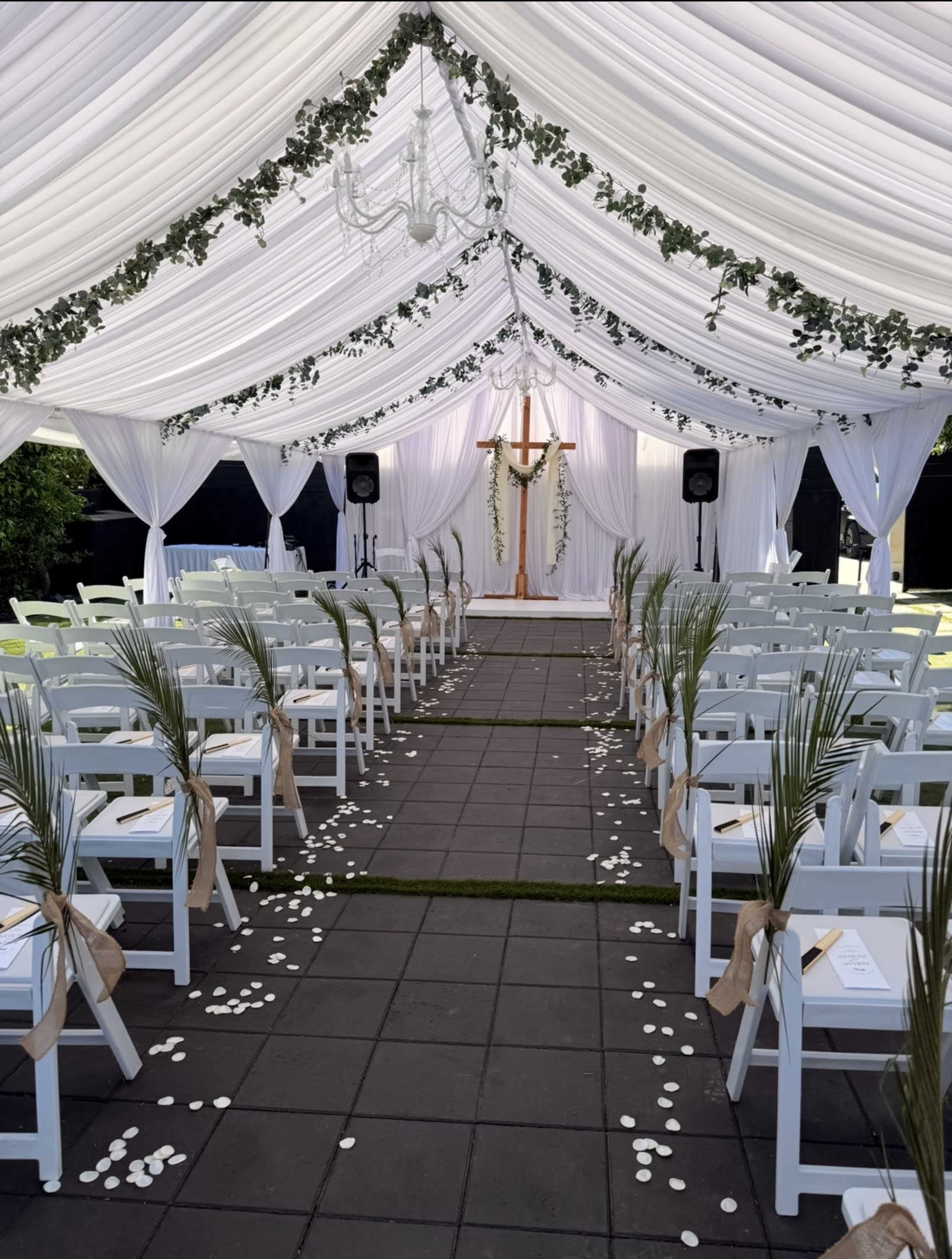 The image shows a decorated outdoor wedding ceremony setup under a white tent, featuring rows of white chairs, a cross with floral accents at the altar, and petals scattered along the aisle.