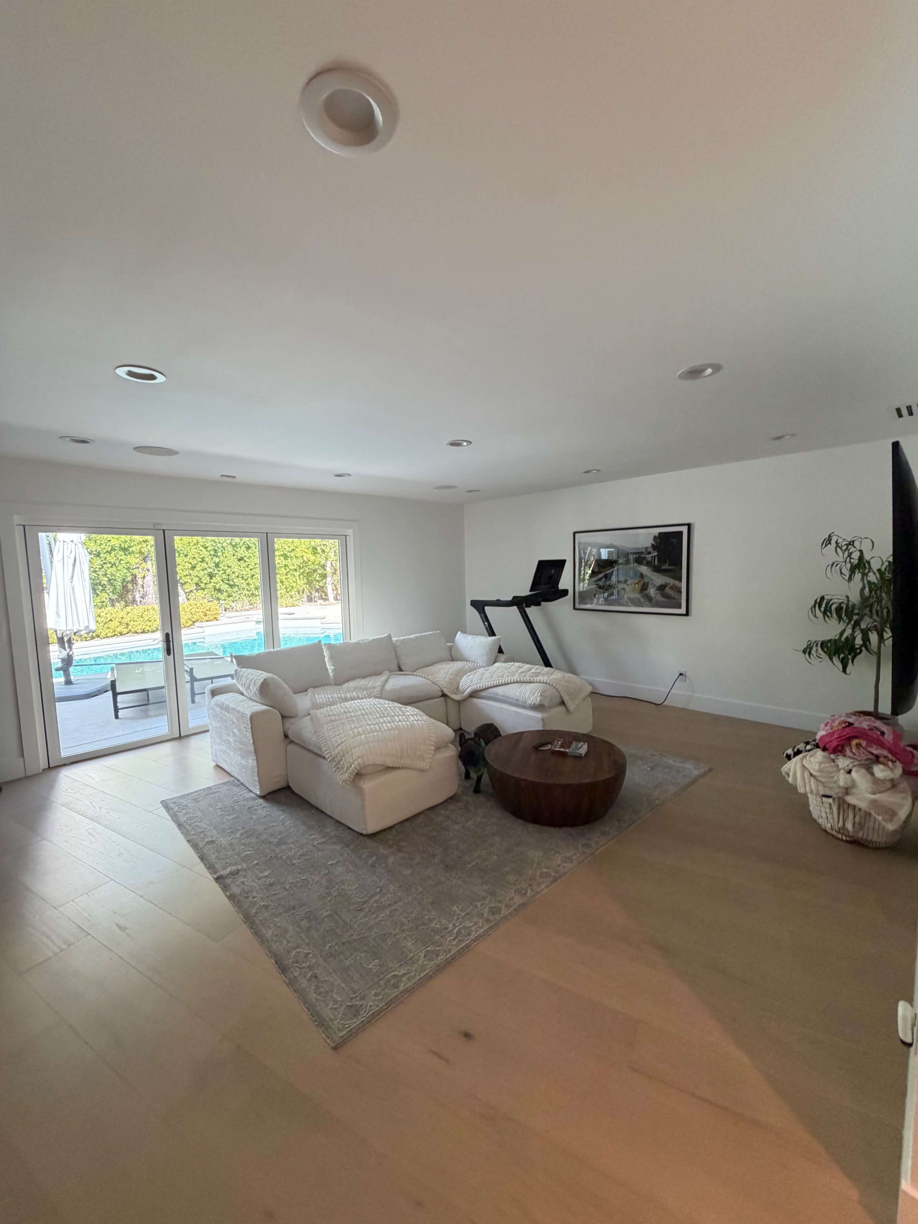 The image shows a modern, airy living room with a white sectional sofa, a wooden coffee table, and a treadmill, overlooking a pool area through large windows.