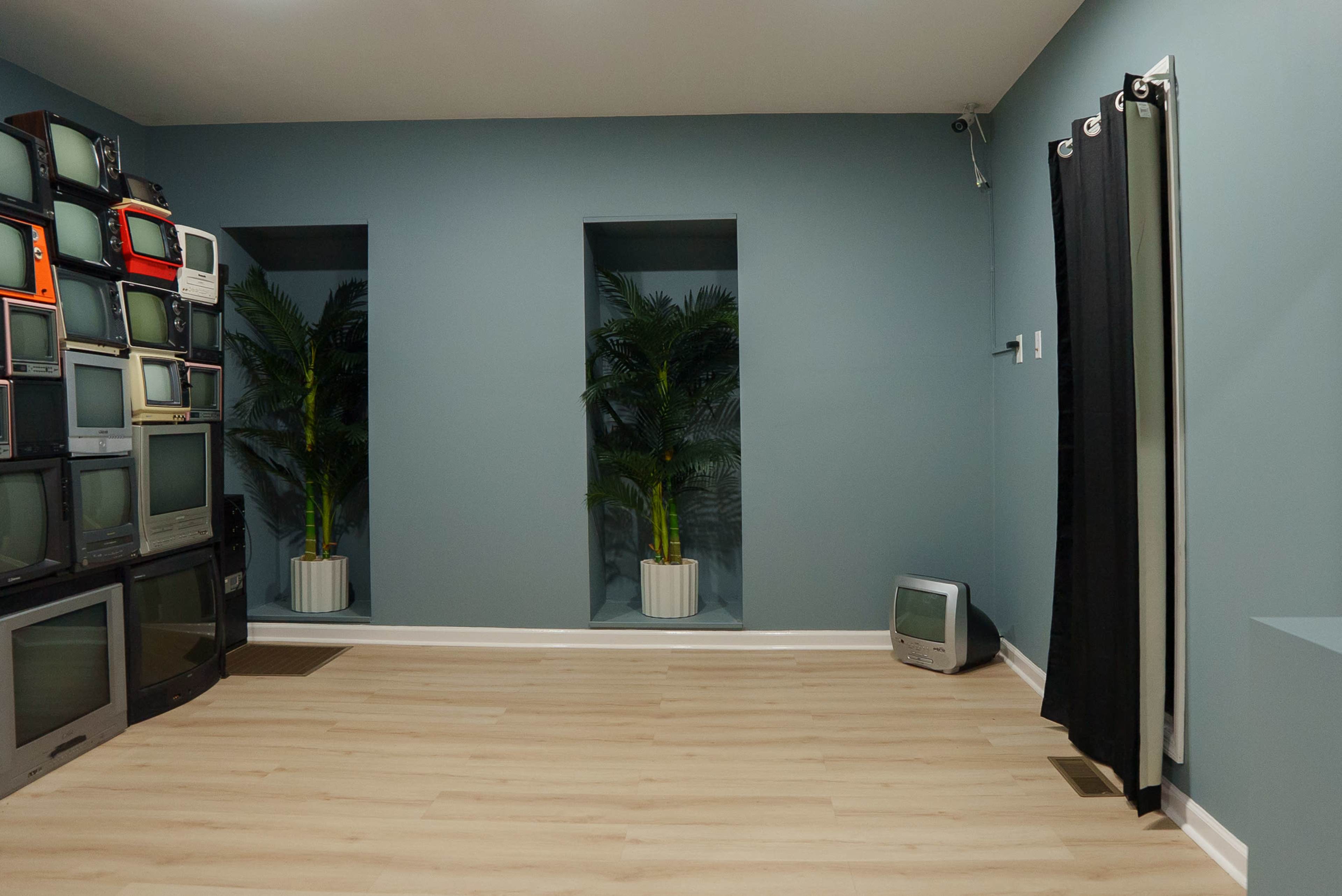 The room features a stack of old television sets on one wall, two window niches with potted plants, and a small TV on the floor.