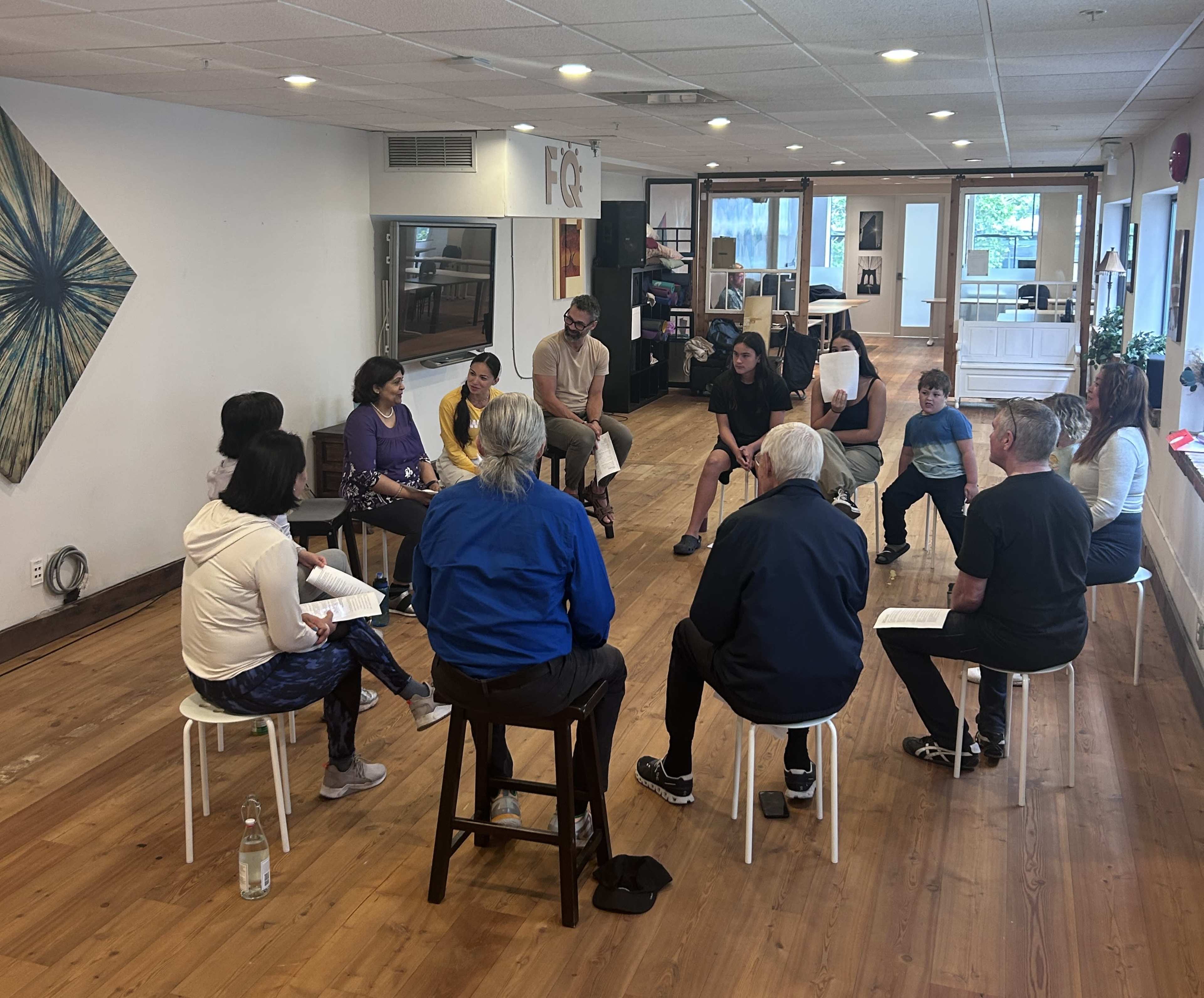 A group of ten people sits in a circular arrangement in a well-lit room with wooden floors, engaged in discussion.