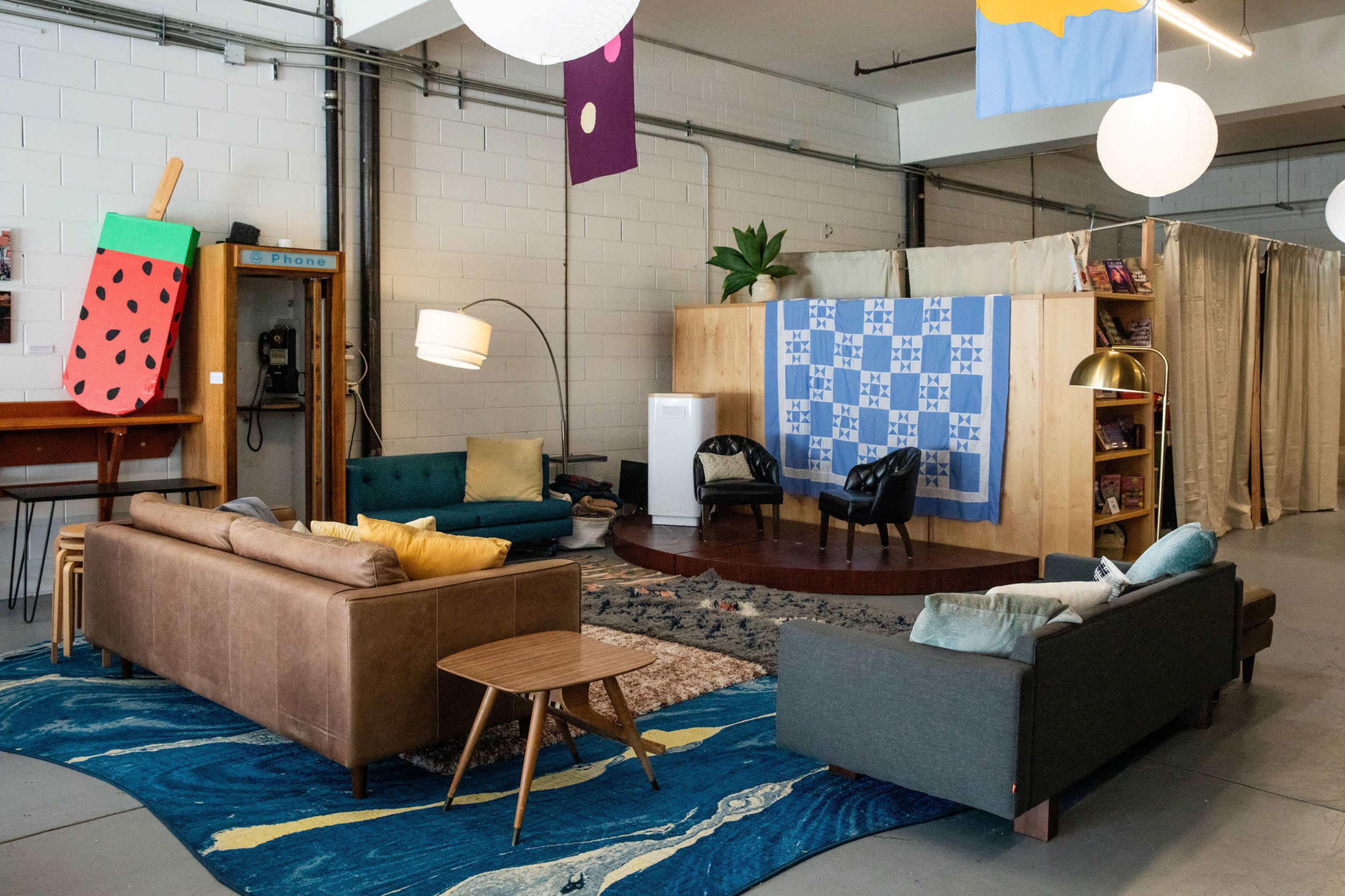 The image shows a modern, open-space seating area featuring two sofas, a coffee table, a patterned rug, and a small stage area with a backdrop and plants.