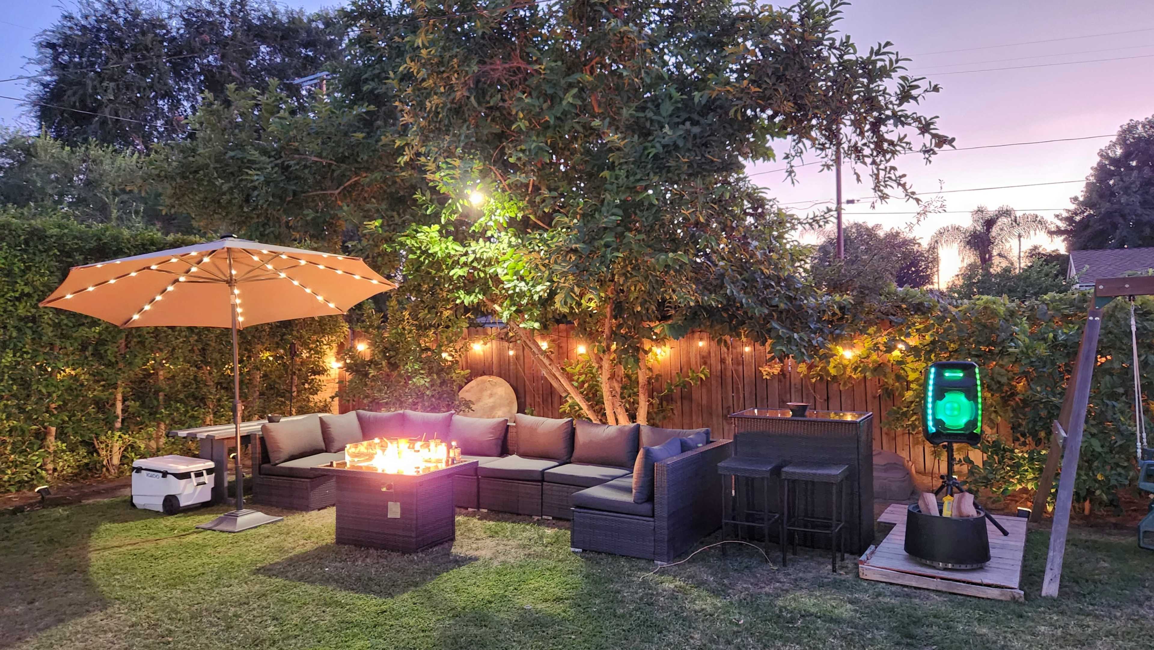 A backyard setting features a fire pit surrounded by a sectional sofa, an umbrella, string lights, and a small cooler.