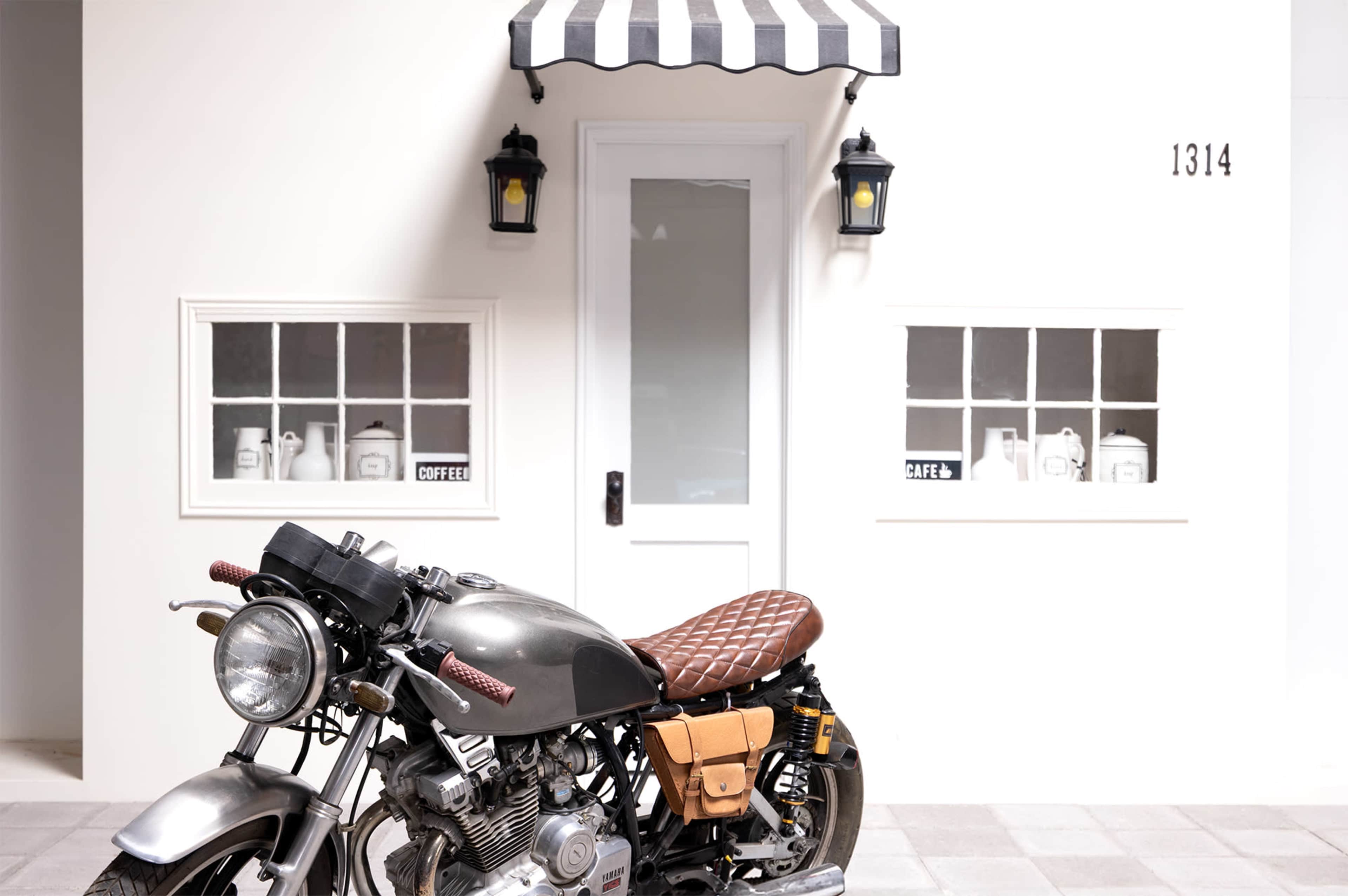 A vintage motorcycle is parked in front of a small café with black and white striped awnings, featuring large windows displaying various ceramic containers.
