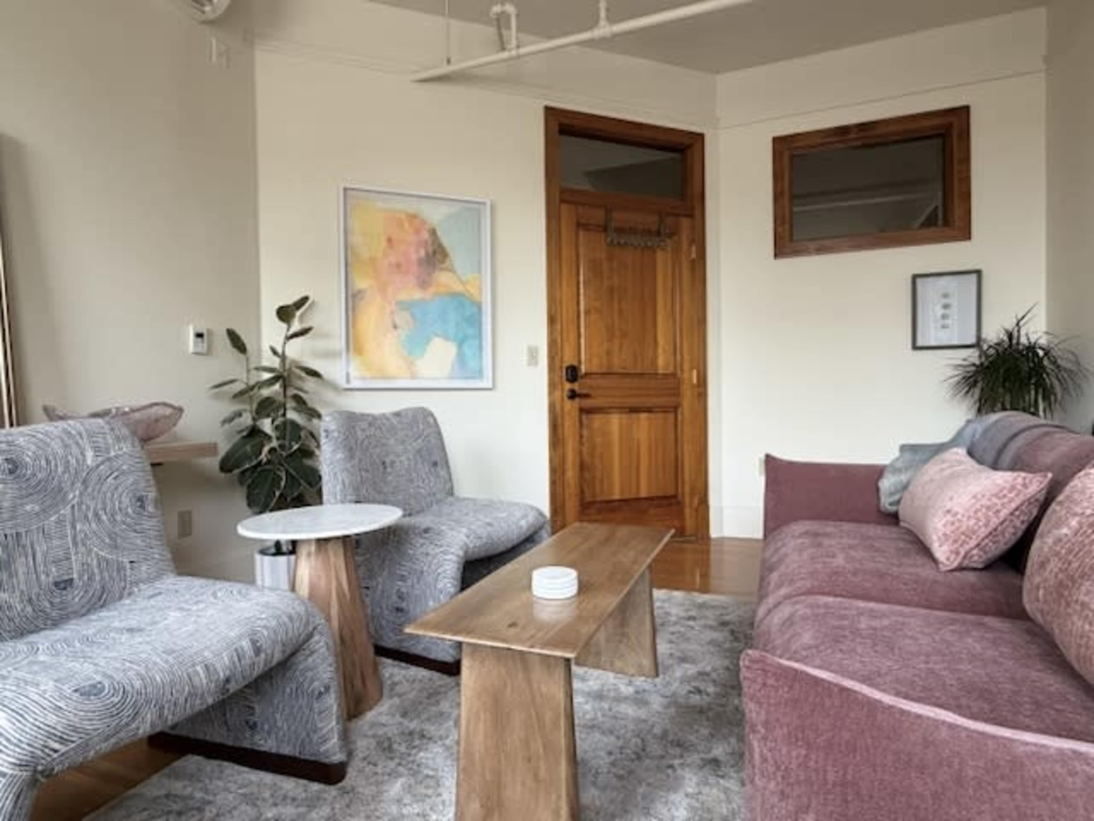 The image depicts a modern living room with two patterned armchairs, a wooden coffee table, and a pink sofa arranged in front of a door and a wall with abstract art.