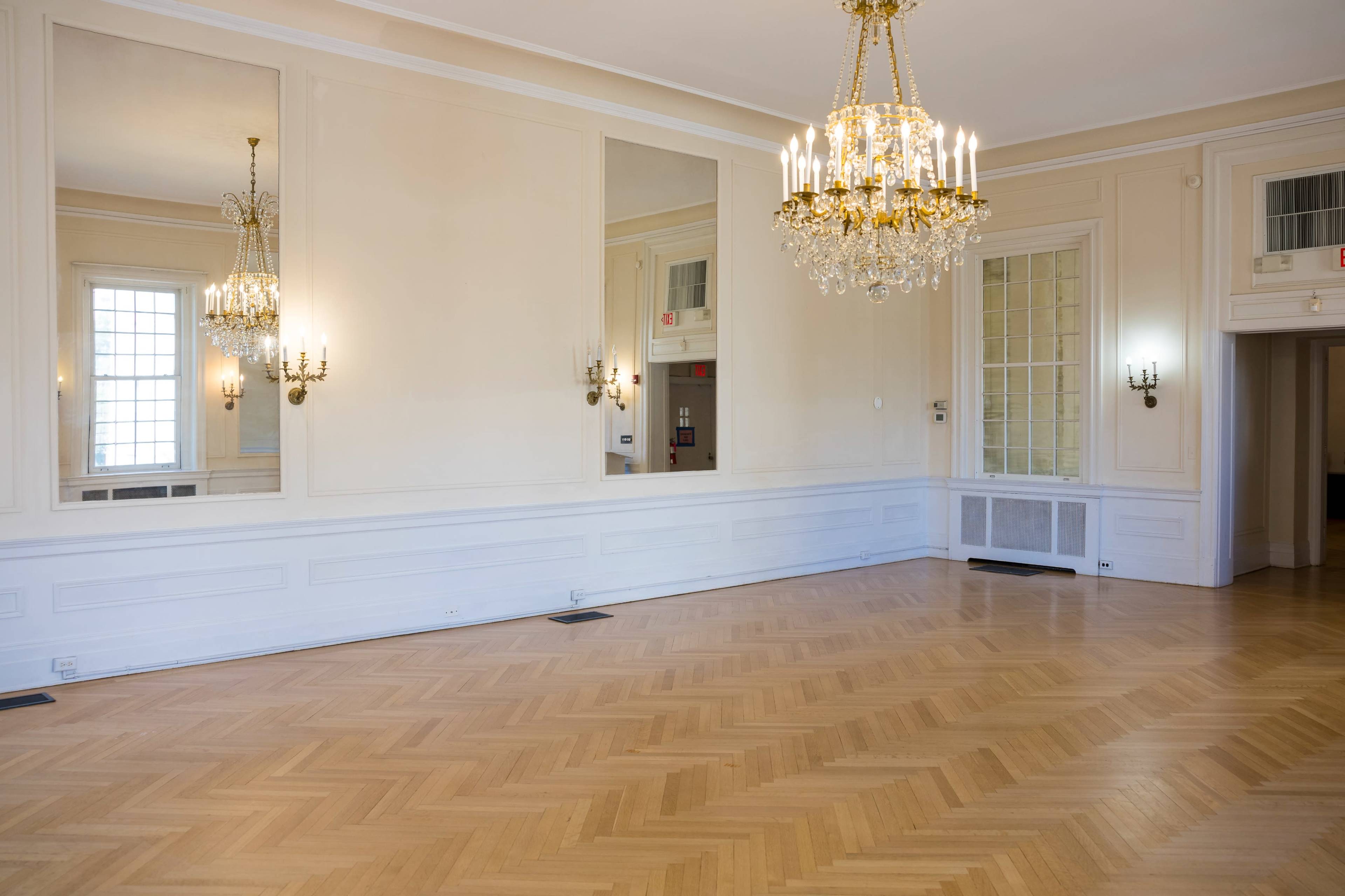 The image shows a spacious room with hardwood flooring, large mirrors on one wall, and a chandelier hanging from the ceiling.
