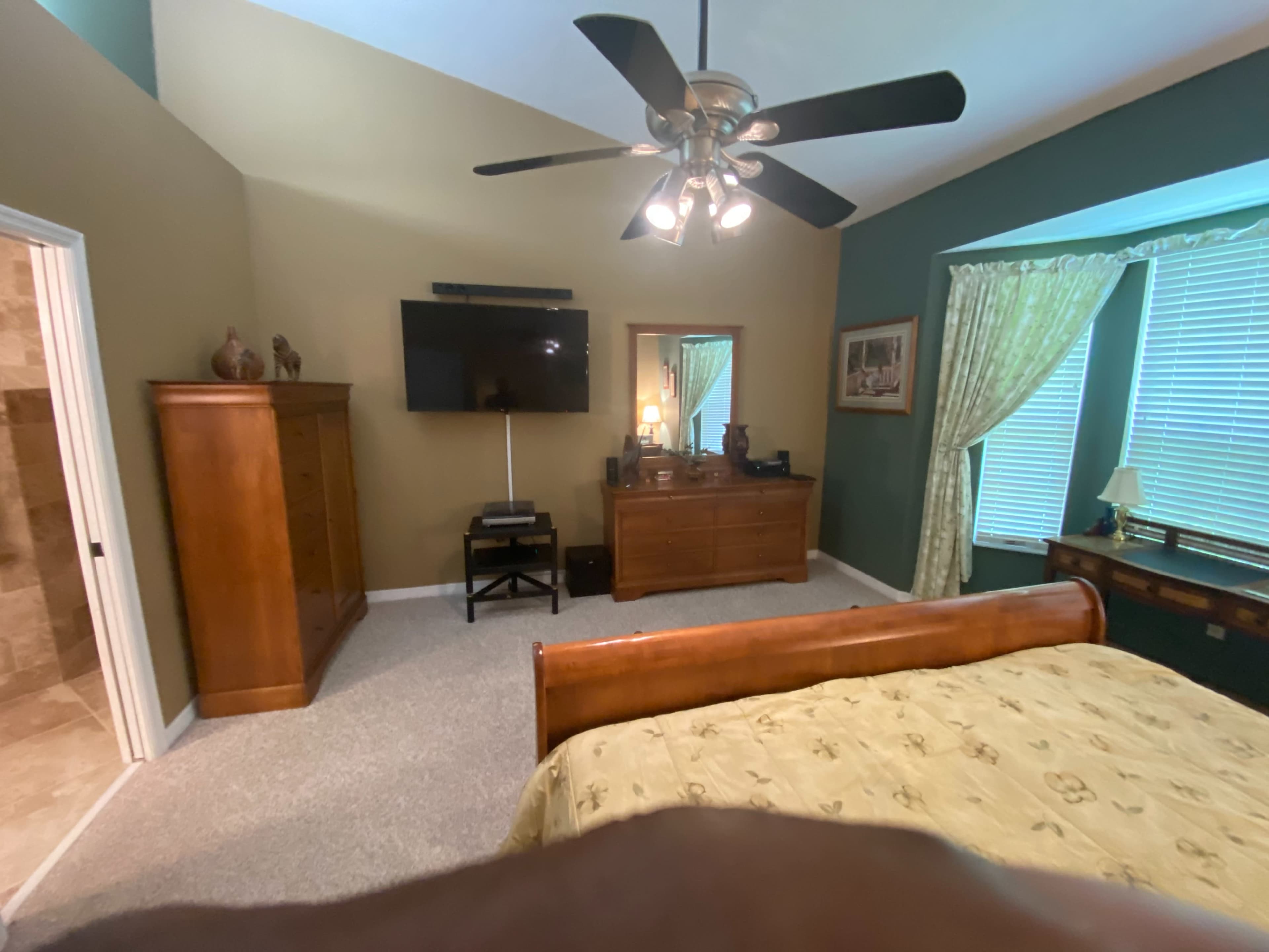 A neatly arranged bedroom features a wooden bed, a dresser, a television mounted on the wall, and a small seating area with a window.