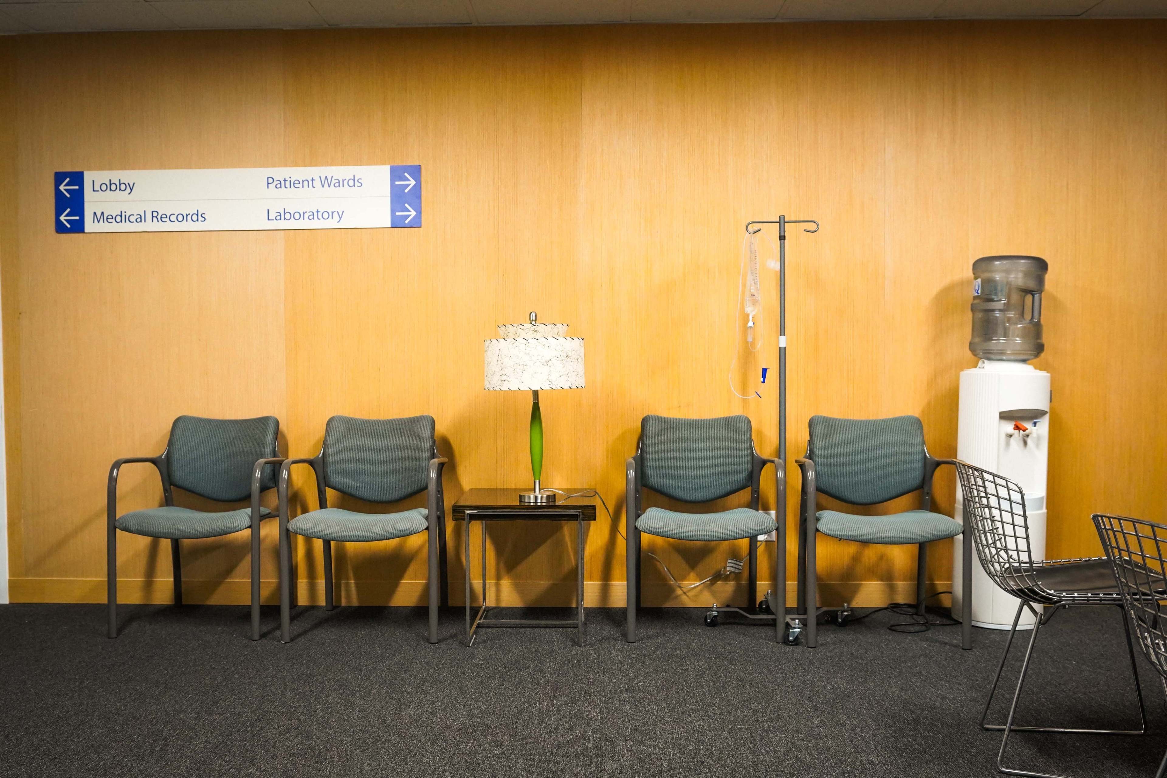 The image shows a waiting area with a row of four chairs, a small table with a lamp, an IV stand, and a water cooler against a wooden wall with directional signs.