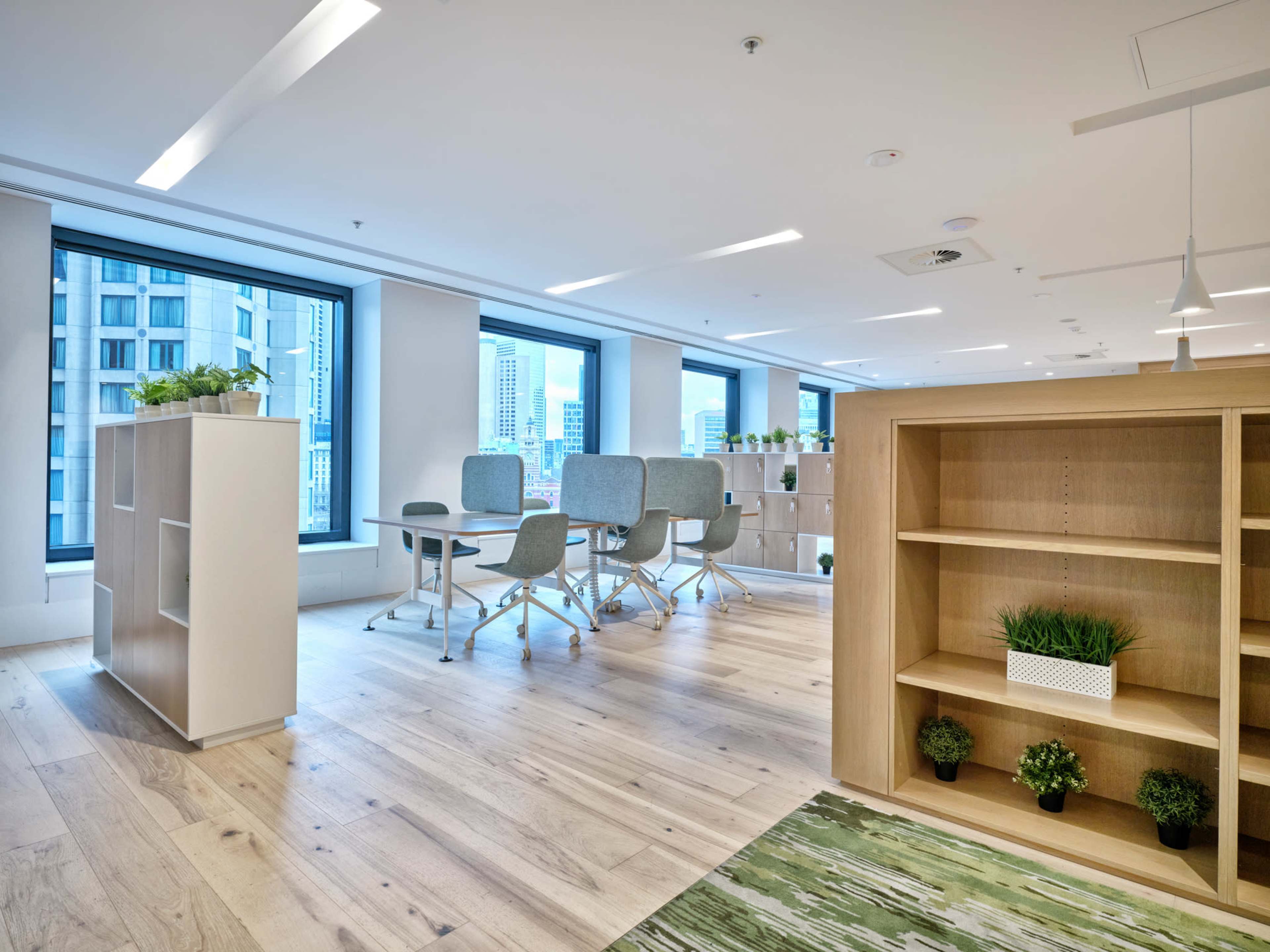 The image shows a modern office space featuring large windows, light wooden flooring, and a mix of seating and work areas with green plants.