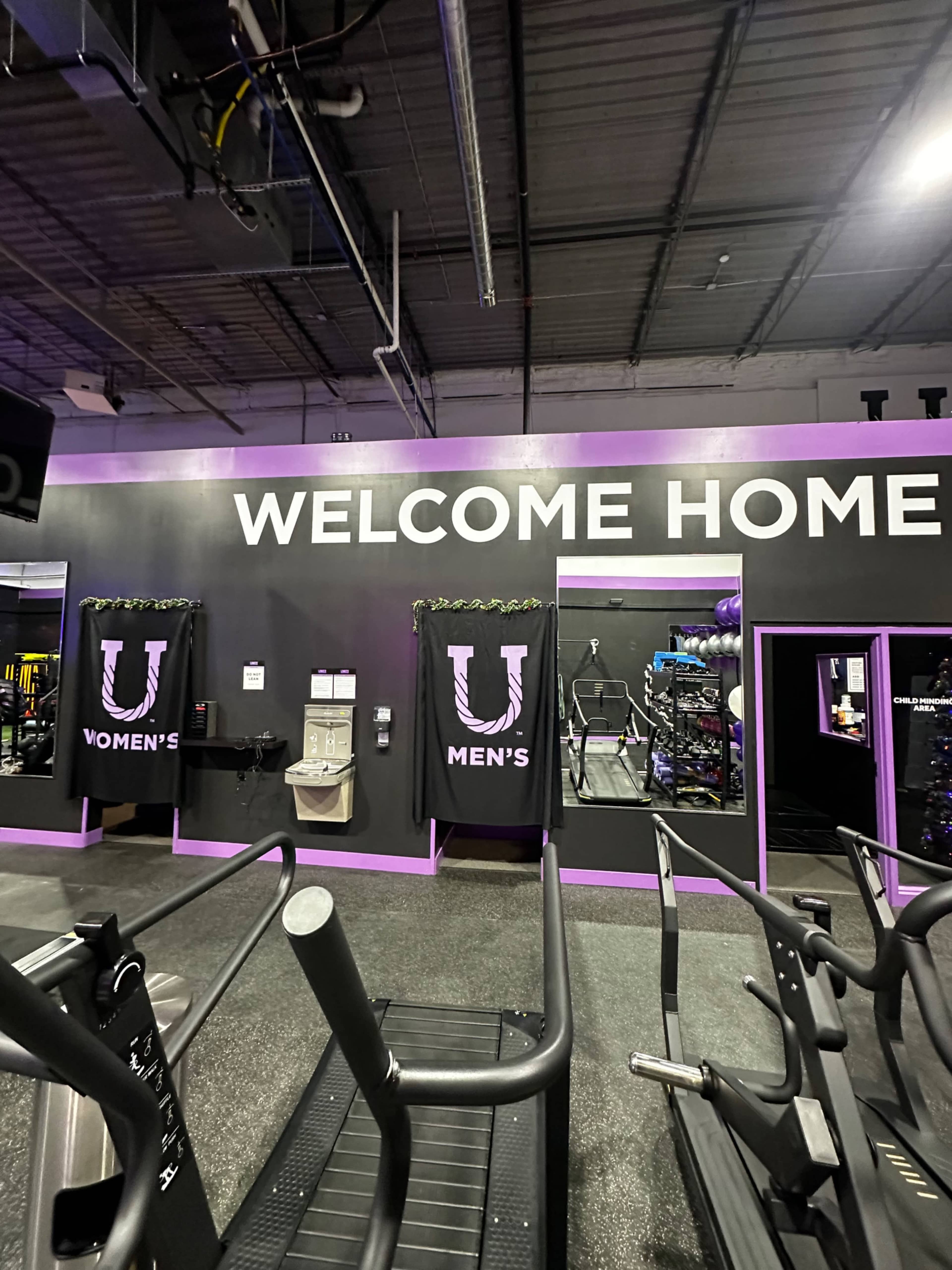 A gym interior with a wall that has "WELCOME HOME" written above two entrances labeled "WOMEN'S" and "MEN'S."