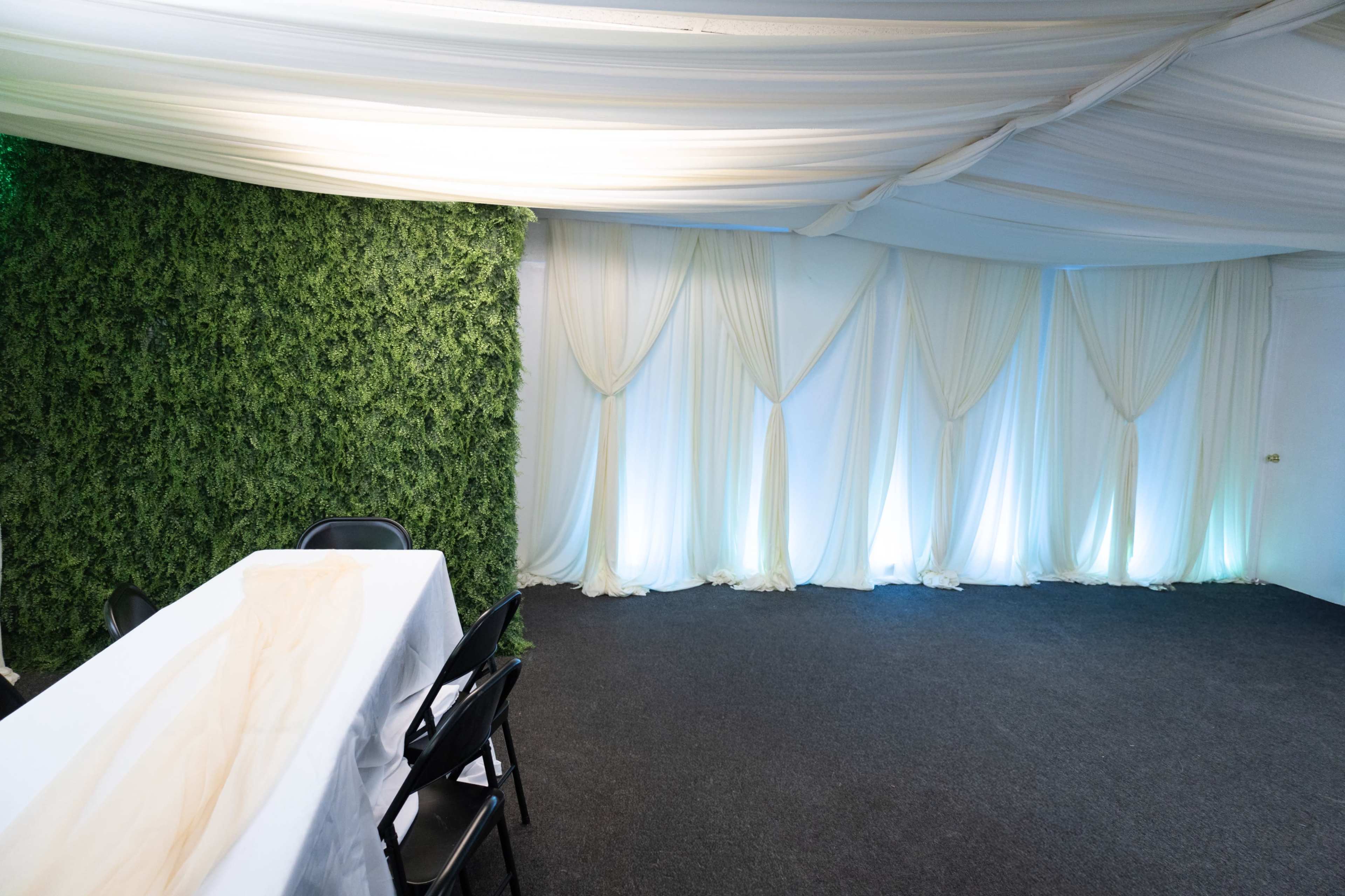 The image shows an indoor event space with a green foliage wall on one side and flowing white drapes along the opposing wall, accompanied by a white table and black chairs.