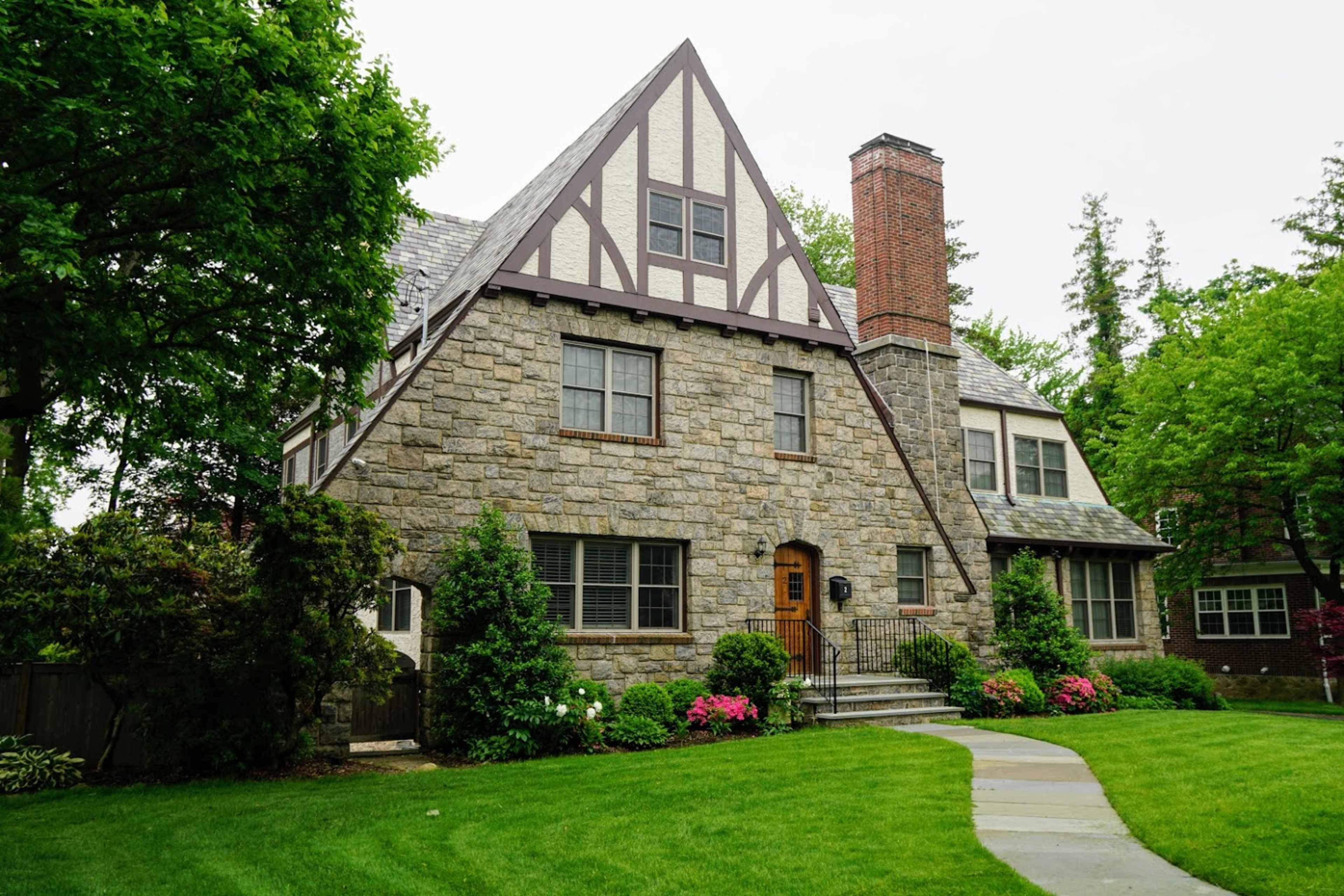 A stone house with a steeply pitched roof and a central wooden door is surrounded by a well-maintained lawn and shrubs.