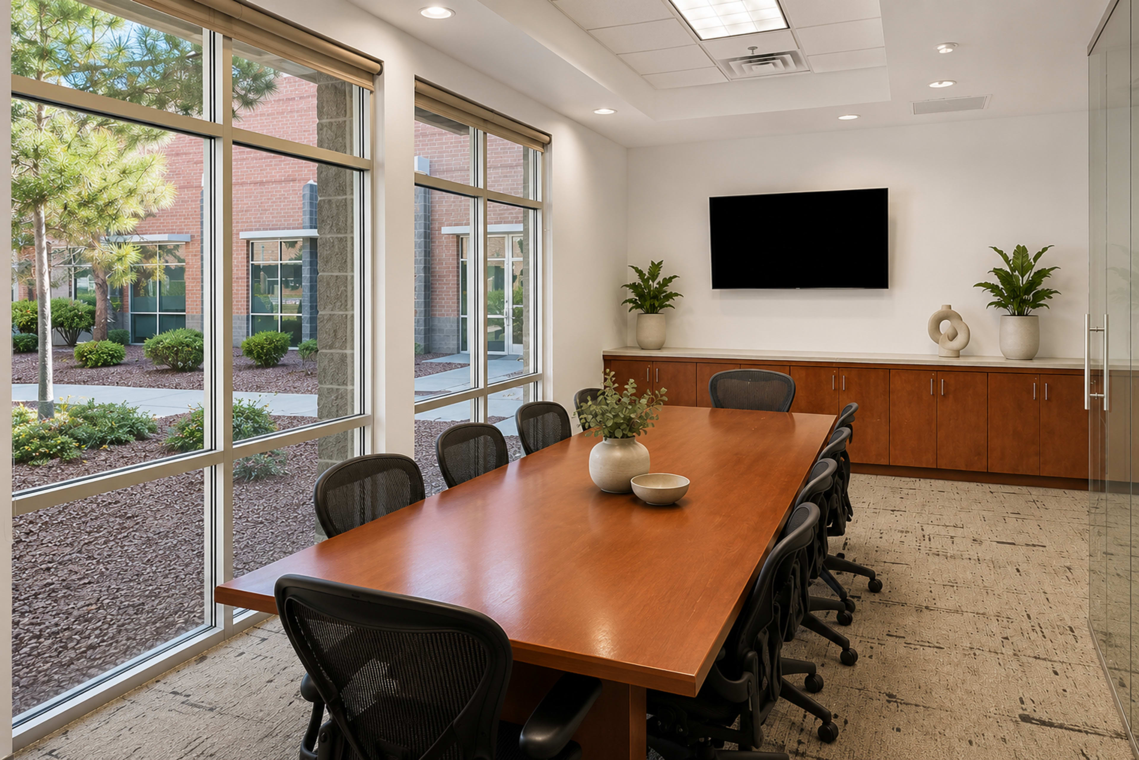 A windowed conference room features a large wooden table surrounded by black chairs, with greenery visible outside.