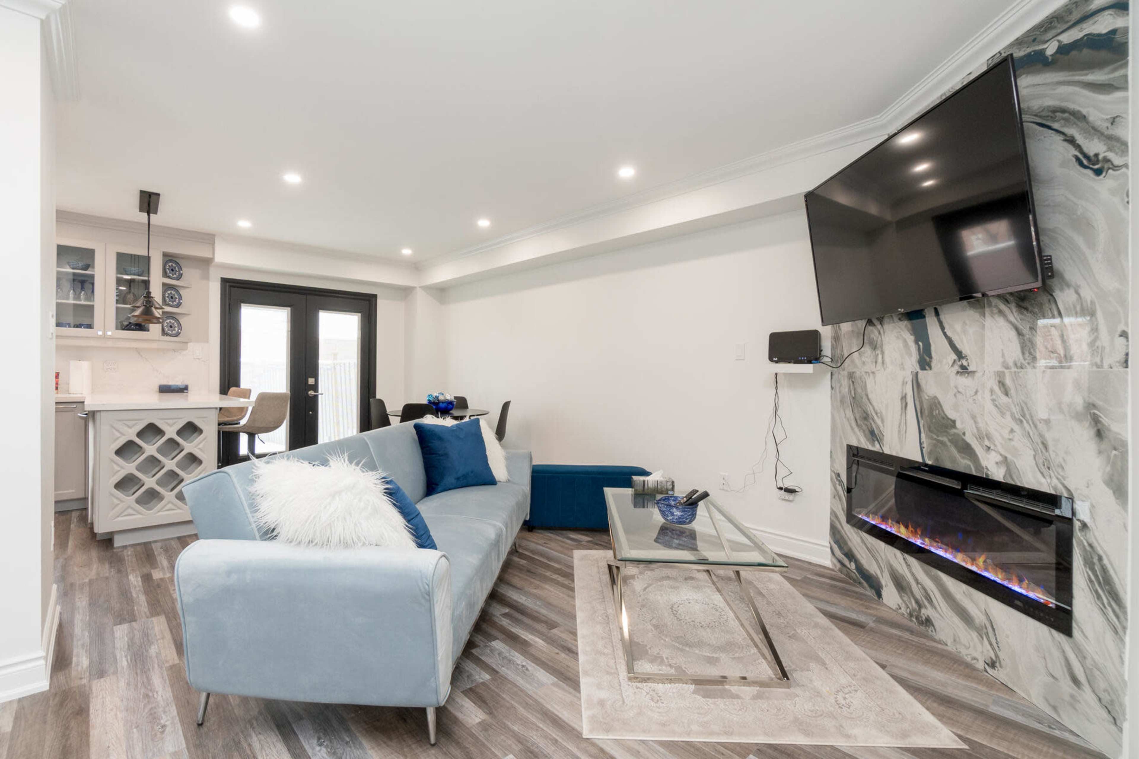 The image shows a modern living room featuring a light blue sofa, a glass coffee table, an electric fireplace with a marbled wall, and a mounted television.