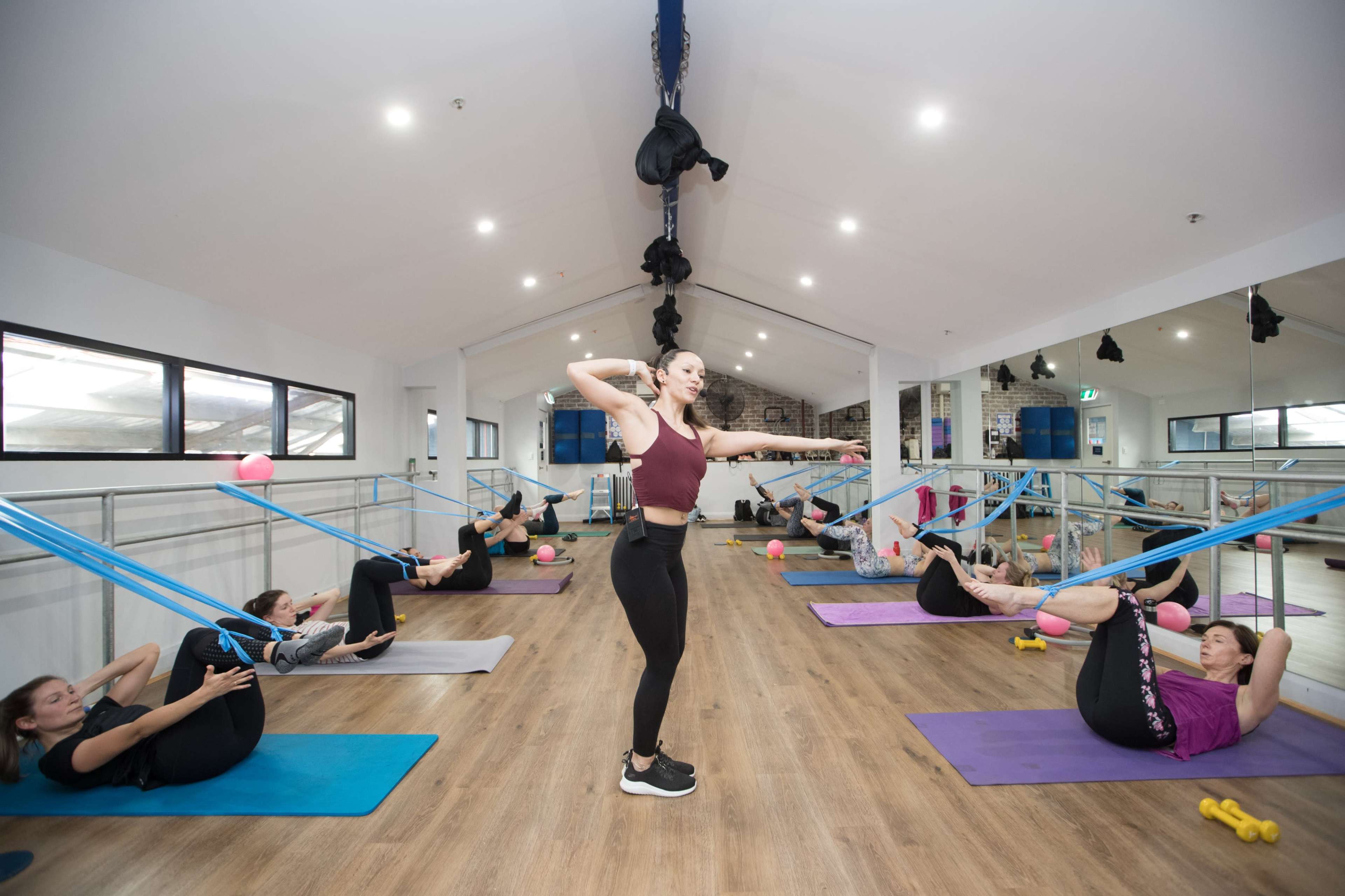 A fitness class is taking place in a studio where participants are using resistance bands while performing exercises on mats.