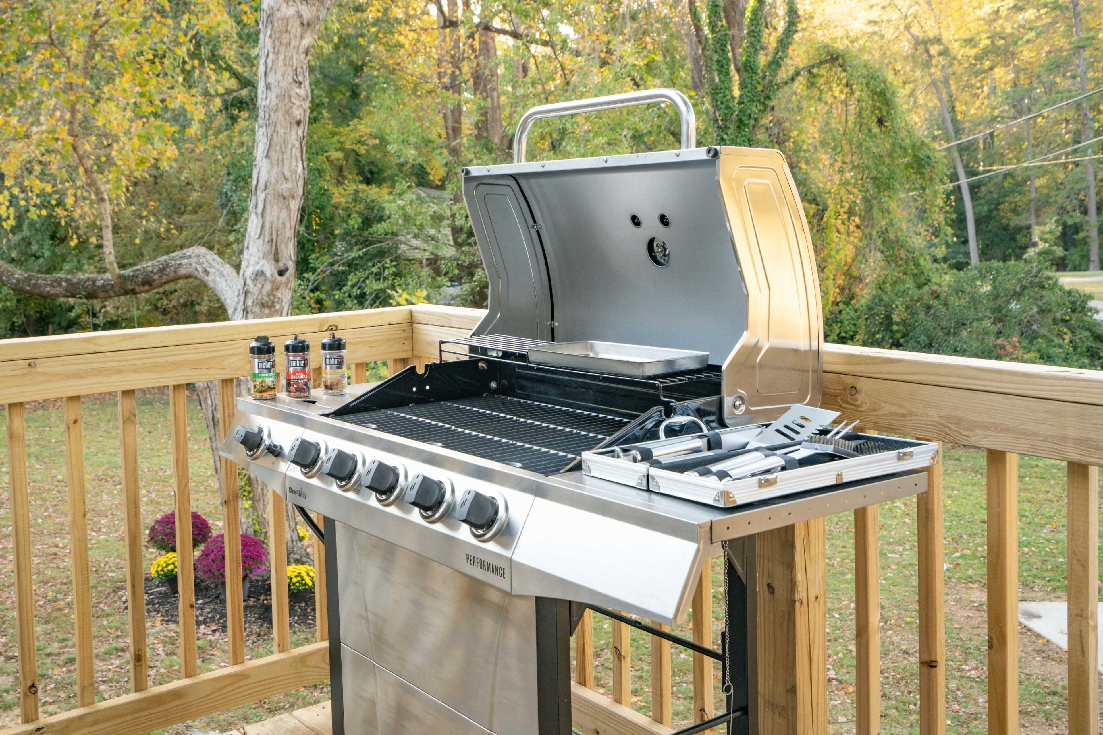 A stainless steel gas grill with the lid open is positioned on a wooden deck, surrounded by trees and colorful flowers.