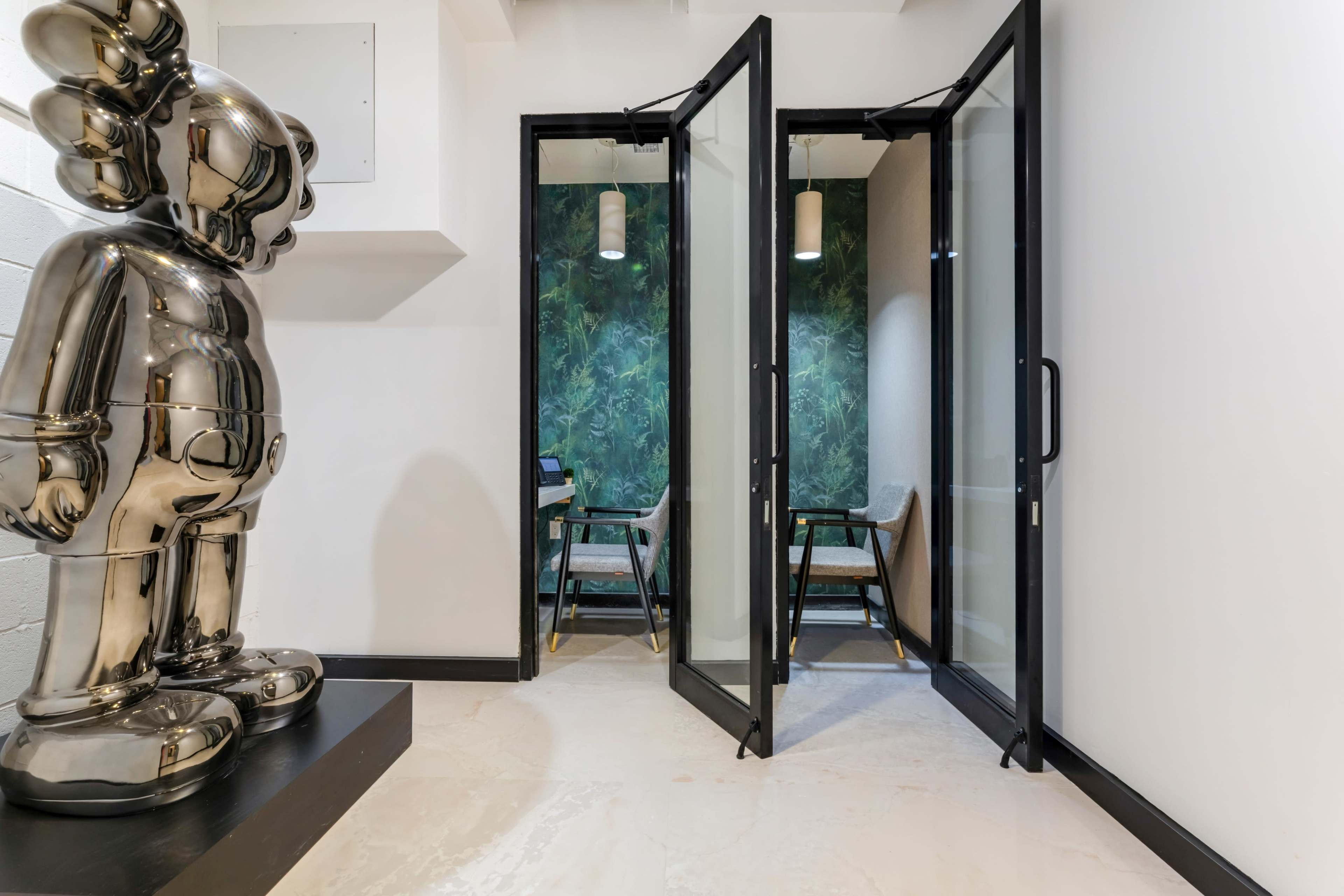 A sleek metallic sculpture stands beside large glass doors that open into a room featuring green foliage wallpaper and two chairs.