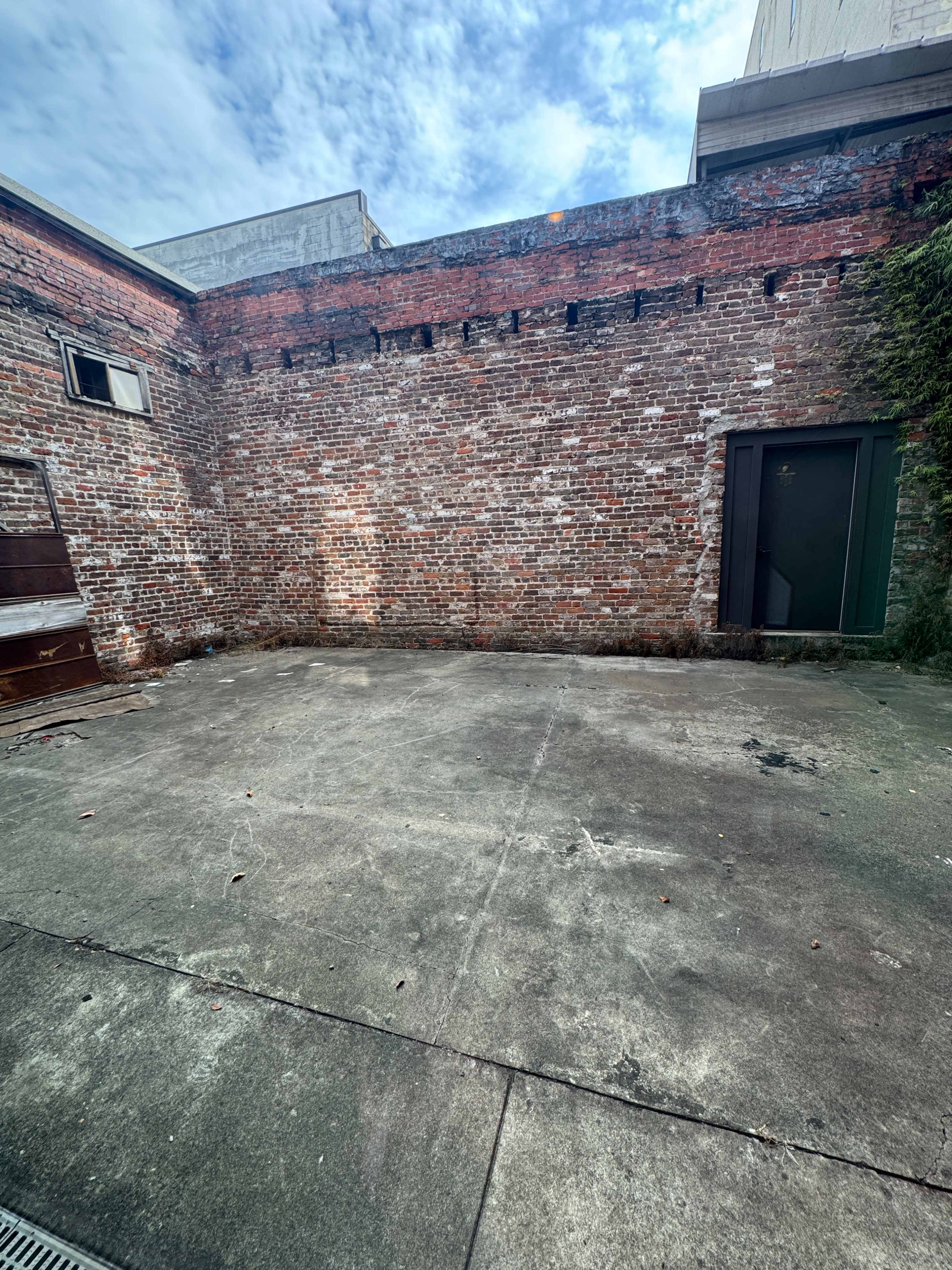The image shows a vacant, concrete courtyard with a brick wall and a door at one end.