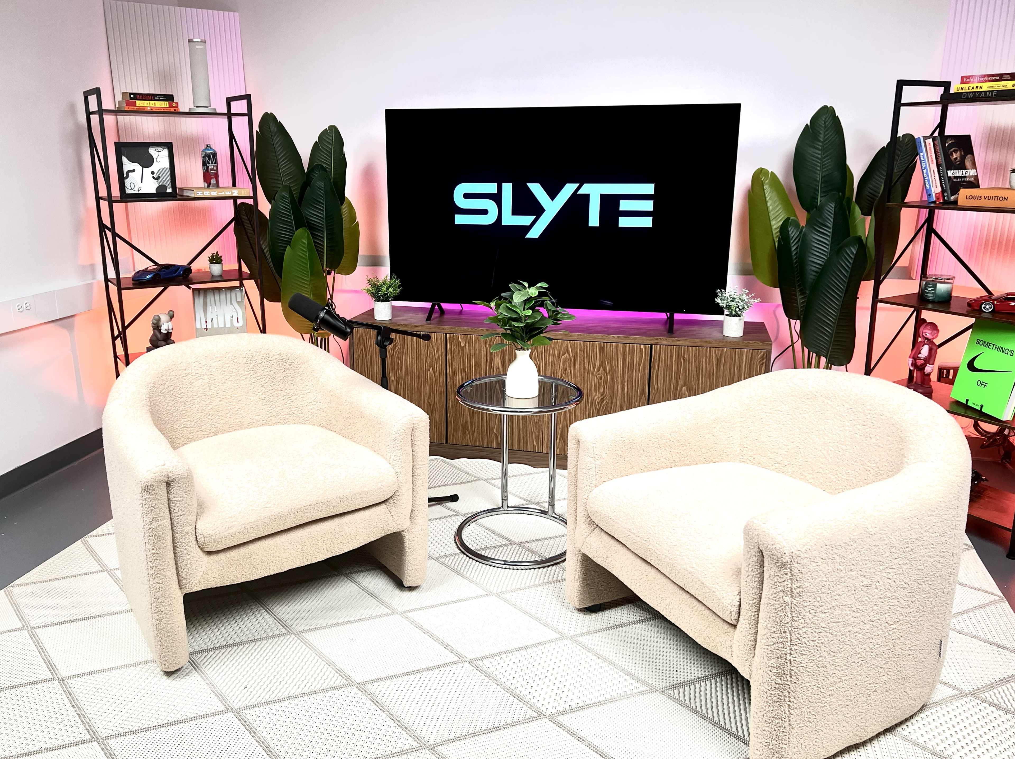 The image shows a modern studio setup with two beige upholstered chairs, a small glass table, a potted plant, and a large screen displaying the word "SLYTE" in front of a softly lit background.