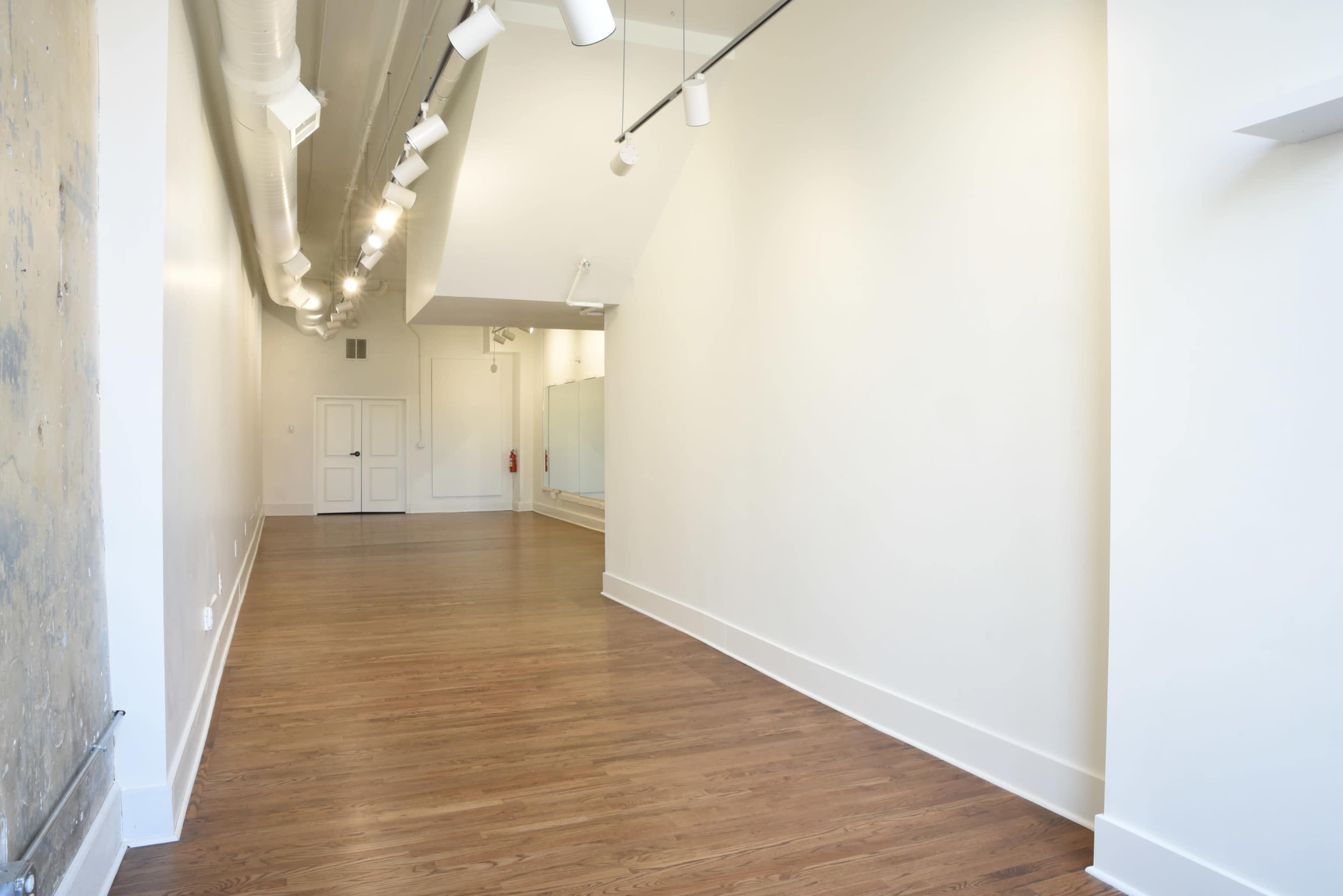 The image shows a large, empty room with hardwood floors, white walls, and track lighting along the ceiling.