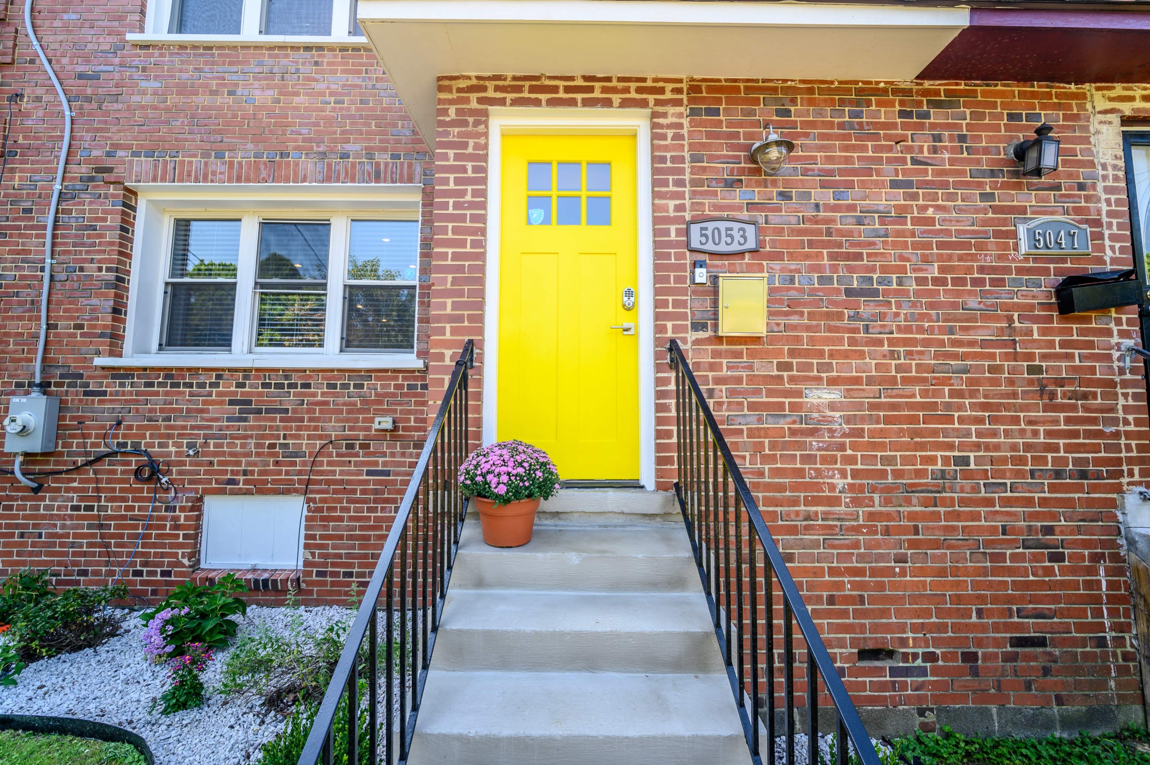 A brick building entrance with a bright yellow door, flanked by potted flowers and a staircase leading up to the door.
